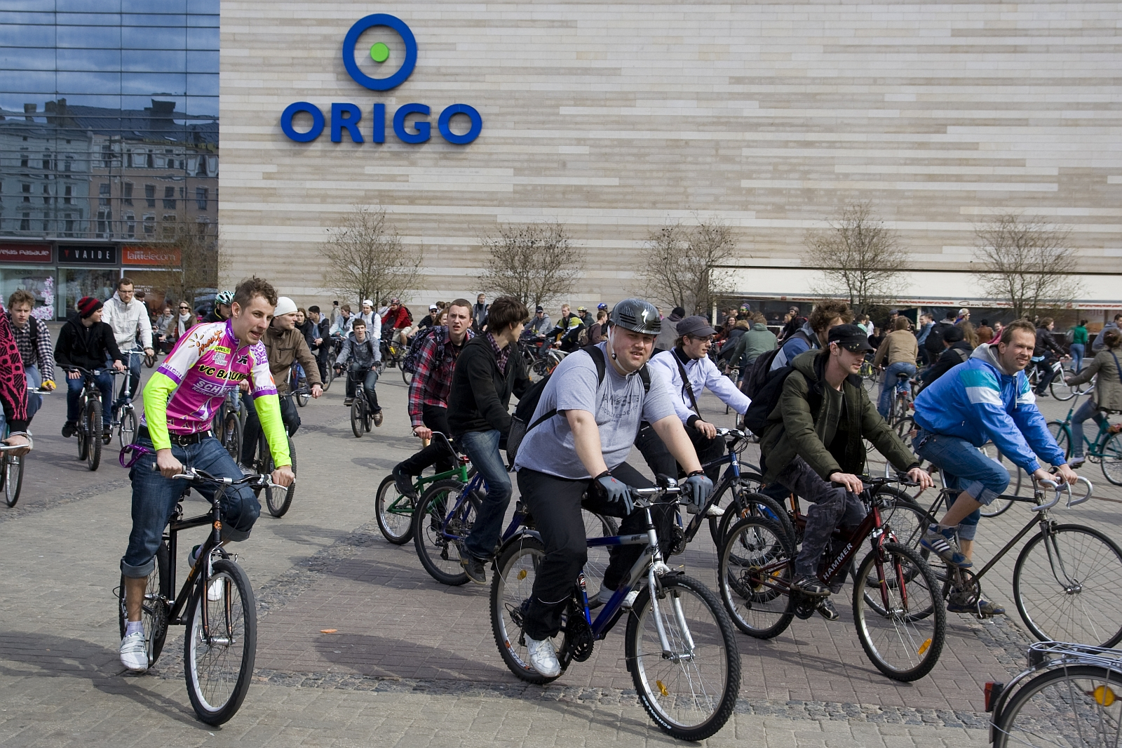 A large group of people participate in a Kritiska masa event, riding bicycles outdoors near a commercial building with the "ORIGO" logo prominently displayed. The cyclists, wearing casual clothing and bike helmets, move in different directions. The background includes other pedestrians, a building wall, and trees.