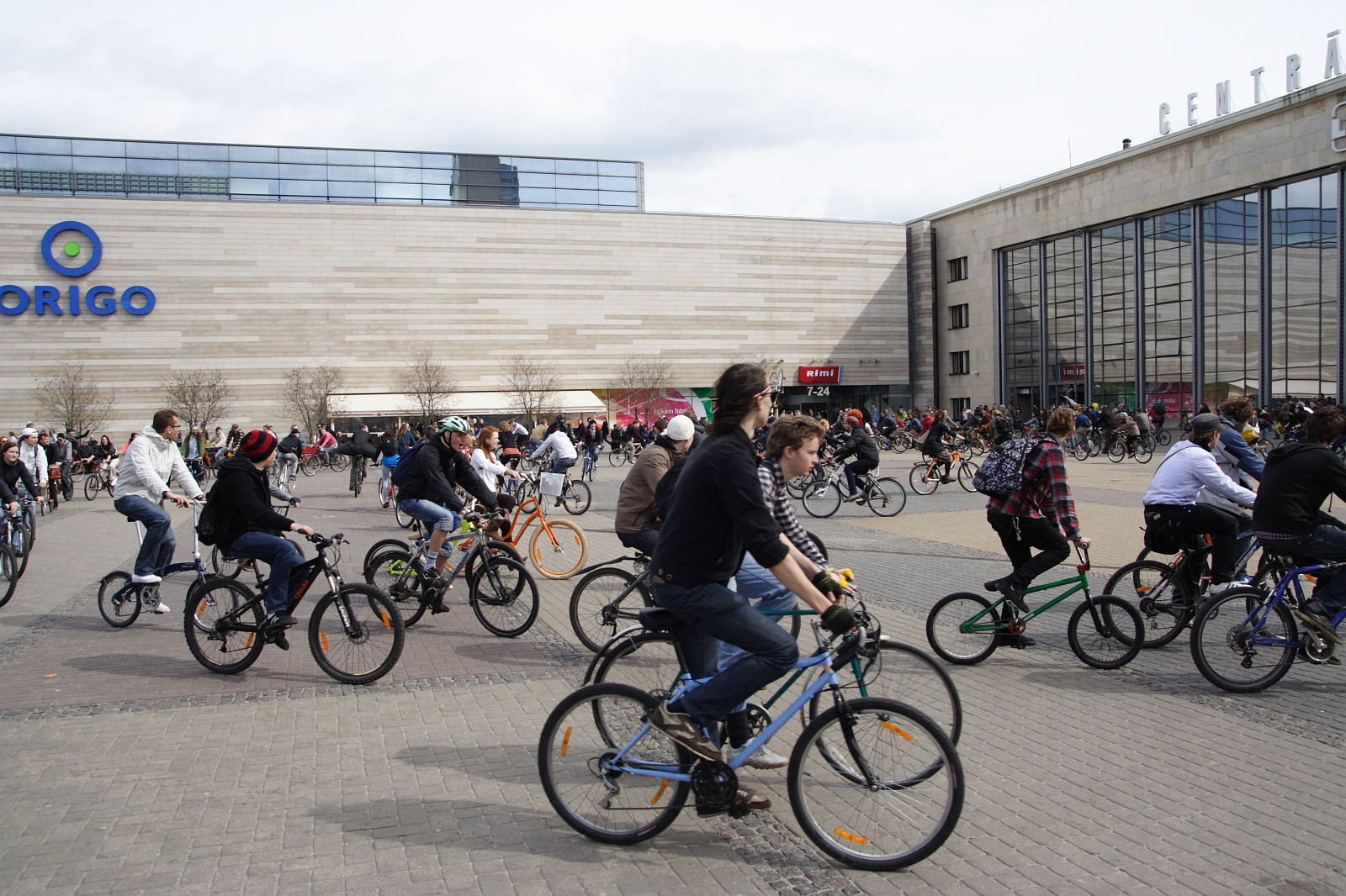 A bustling scene of numerous people riding bicycles in an urban square depicts the Kritiska masa event of 2010. The background shows a modern, large building labeled "Origo." The crowd is diverse, including adults and children with varied bike styles. The sky is overcast, and the atmosphere appears lively and active.