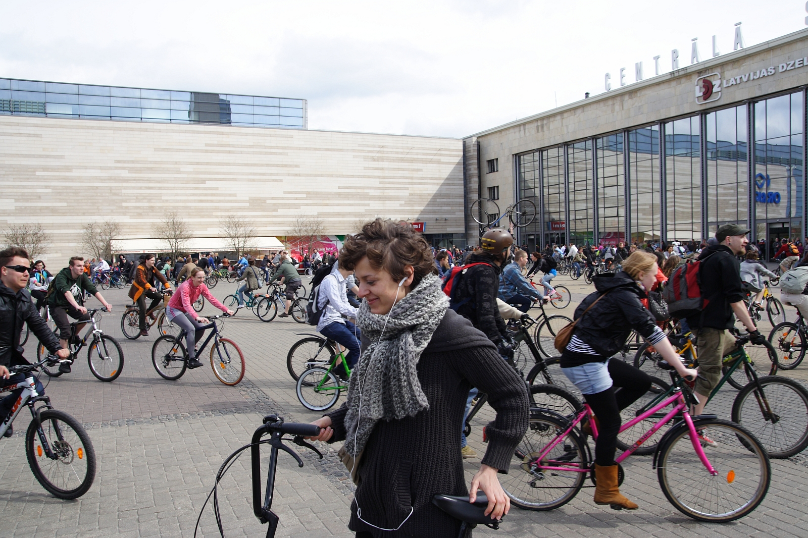 A bustling urban square filled with numerous cyclists riding in different directions. In the foreground, a woman in a gray scarf and dark jacket is walking her bicycle with a smile on her face. Behind her, many people are also on bicycles, reminiscent of the Kritiska masa event from 2010, with buildings visible in the background.