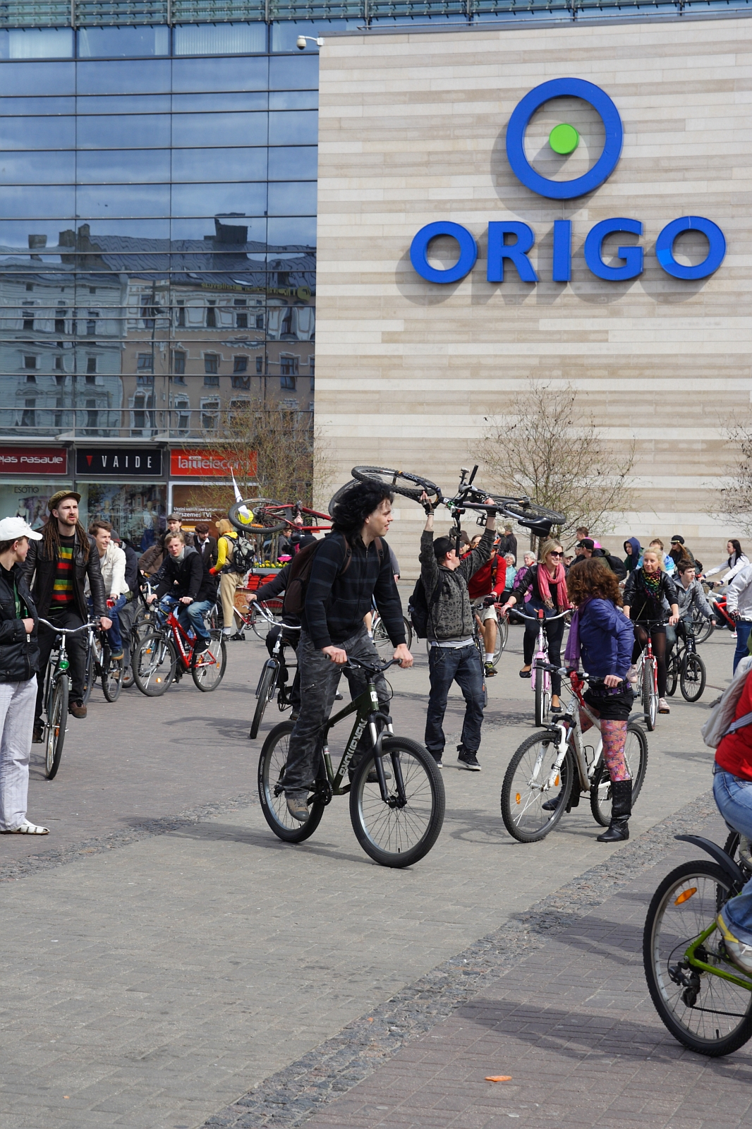 A large group of people on bicycles gathered in front of a modern building with a sign that reads "ORIGO." Reminiscent of the 2010 Kritiska masa events, the cyclists are of various ages, wearing casual clothing. The area is paved with riders and those standing with their bikes. Reflective windows show additional buildings.
