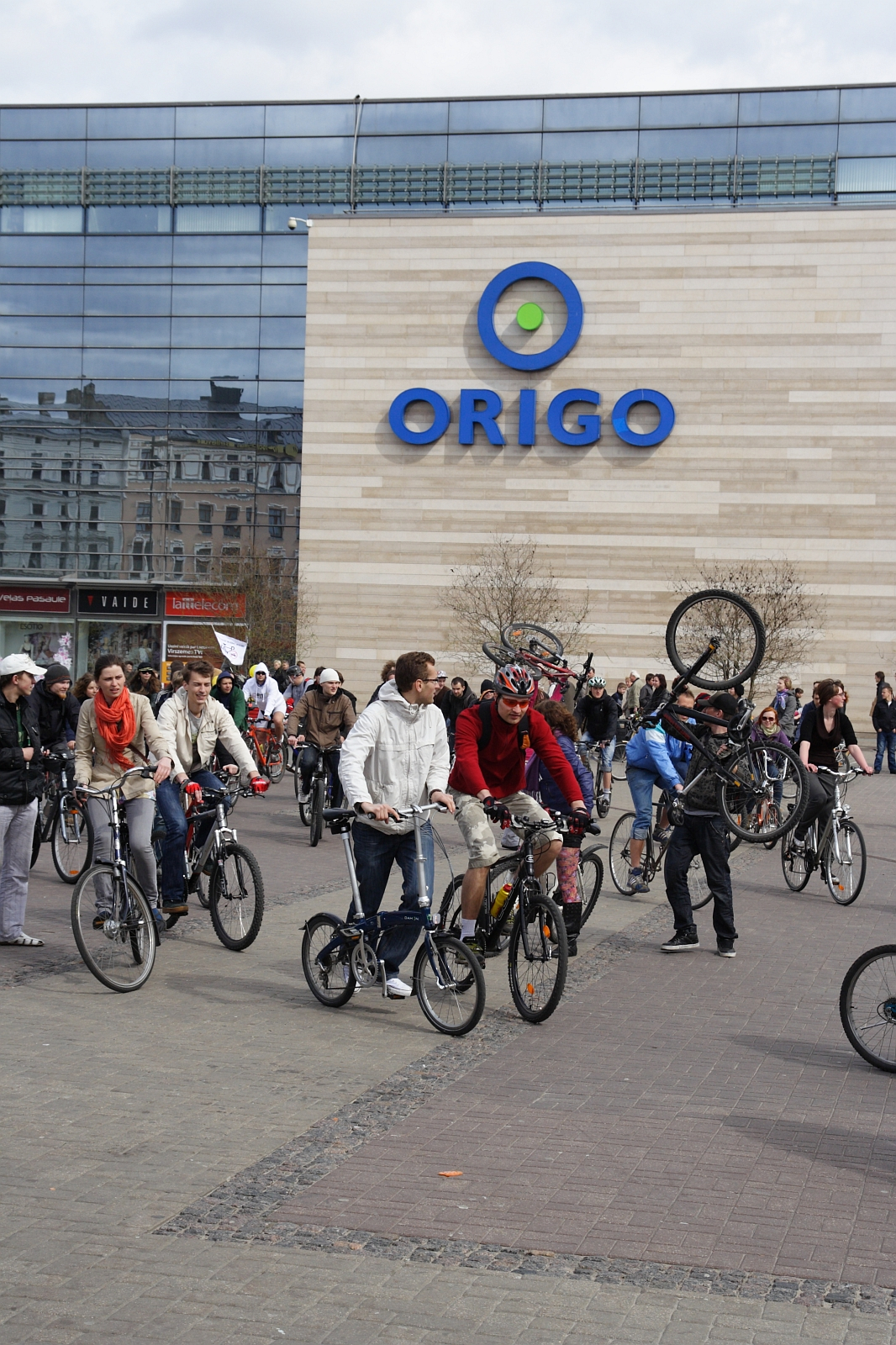 A group of cyclists is gathered in front of the Origo shopping center, reminiscent of the Kritiska Masa event in 2010. Some individuals are performing stunts, including a person mid-wheelie. Numerous spectators are watching the event. The building features a large glass facade and beige walls with the blue "ORIGO" logo prominently displayed.