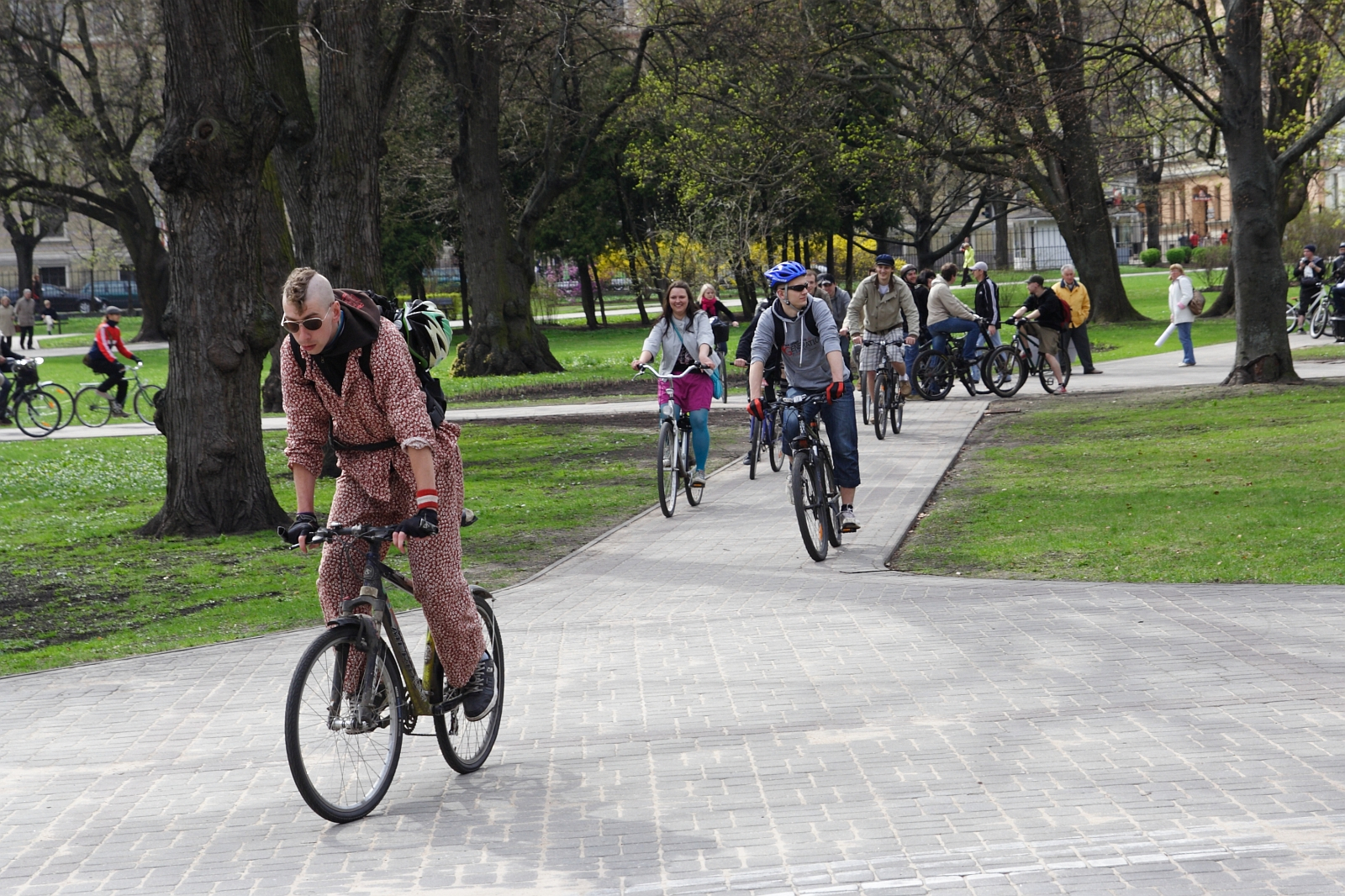 A cyclist wearing a patterned onesie and sunglasses rides along a paved path in a park. Close behind him are several other cyclists, including a woman in a pink jacket and a man in a blue helmet. The scene resembles the 2010 Kritiska masa event, with people on bikes surrounded by green grass and trees.
