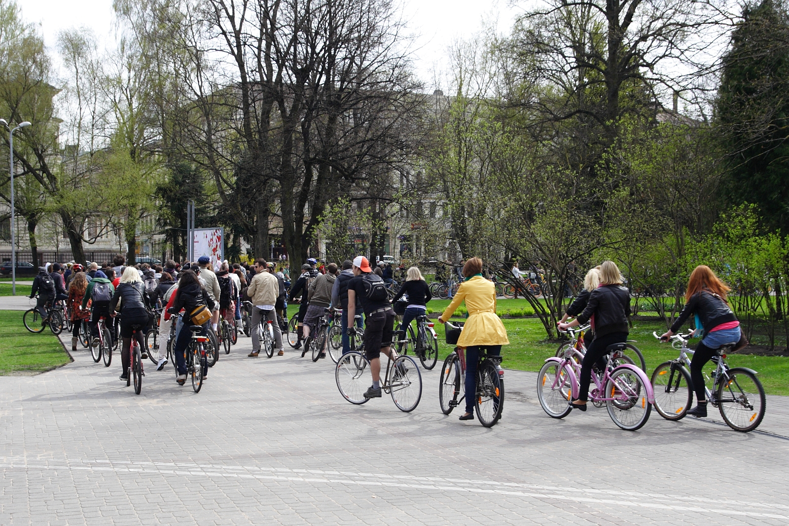 A group of people riding bicycles on a paved path in a park surrounded by trees with budding leaves. The cyclists, dressed in various casual outfits, evoke the spirit of a 2010 Kritiska masa event or leisurely group ride. Buildings are partially obscured by the trees in the background.