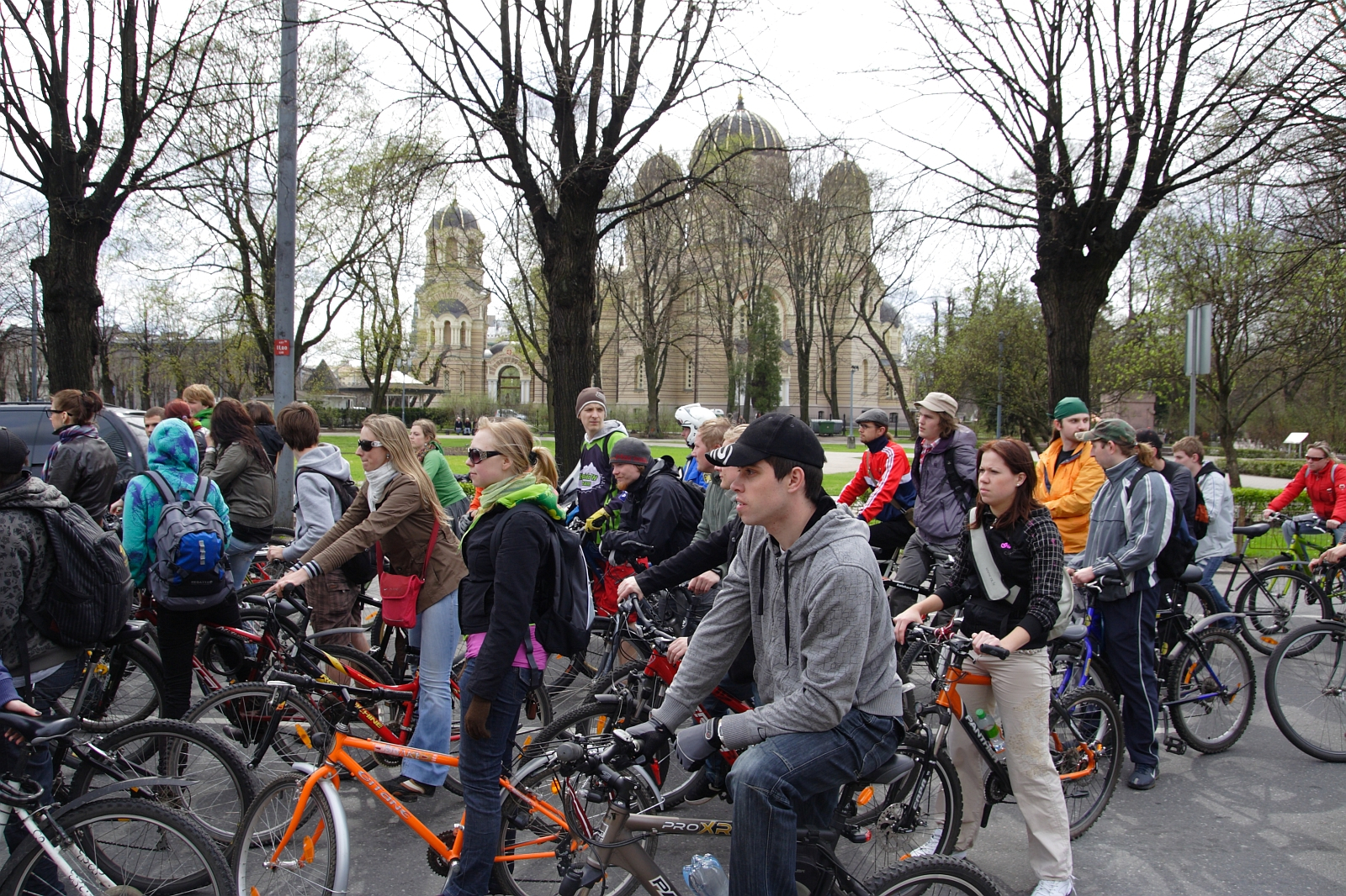 A large group of people on bicycles is gathered on a street for the Kritiska masa event. Dressed in jackets and hoodies appropriate for cool weather, they stand amid bare trees lining the street. In the background, a historic building with domes and arches is visible. The mood is calm and organized, reminiscent of 2010 events.