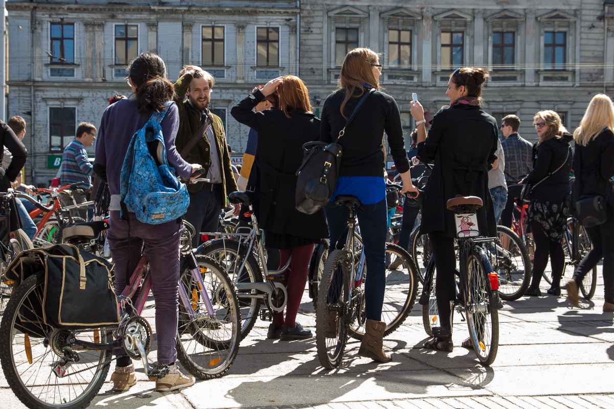 A group of people gathered outdoors with their bicycles in front of an old building during the 2013 Kritiskā masa event. Several individuals are engaged in conversation, and some have their backs turned, displaying colorful backpacks. The weather is sunny, casting shadows on the pavement.