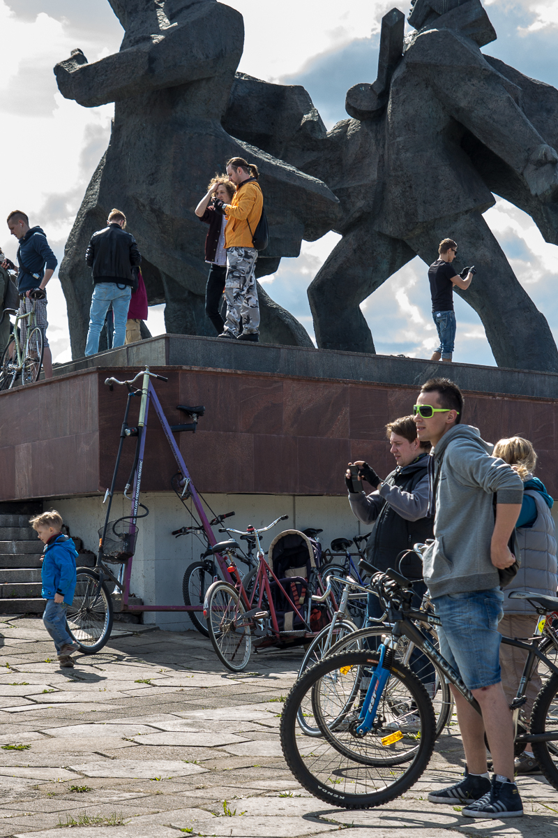 A group of people gathers around large, abstract sculptures. Several are taking photos, and some are riding or standing with bicycles. A tall, unusually designed bicycle stands out. The scene is casual, reminiscent of Kritiskā masa 2013, with individuals of various ages, including children, engaged in leisure activities.