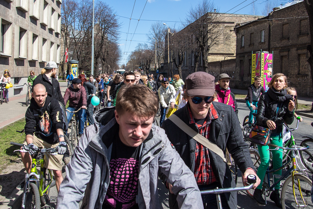A bustling crowd of cyclists pedals down a city street lined with bare trees and brick buildings. The riders, clad in various outfits including jackets and hats, navigate the narrow road together. Colorful signs and street art add vibrancy to the scene under a clear blue sky during Kritiskā masa.