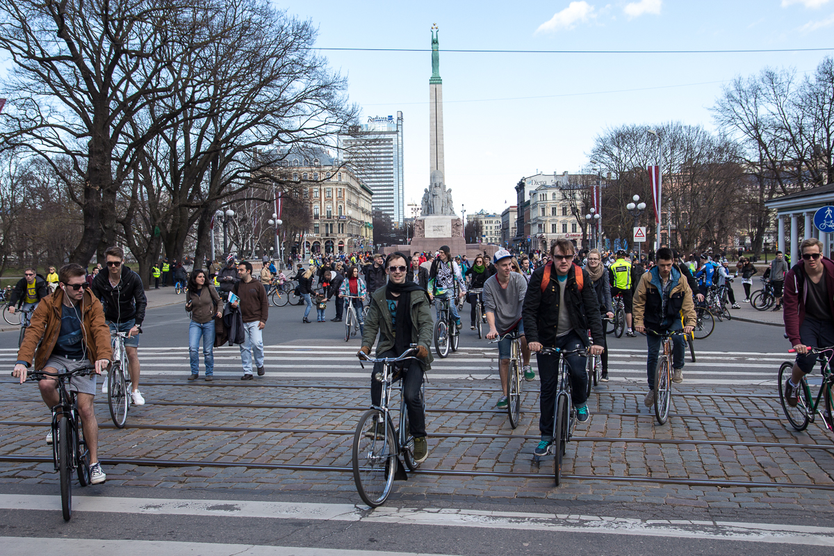 A large group of cyclists rides on a cobblestone street near a monument with a statue on top. Leafless trees and buildings surround the area under a clear sky. Some cyclists wear sunglasses and backpacks, while pedestrians are seen in the background. This 2013 event is part of Kritiskā masa.