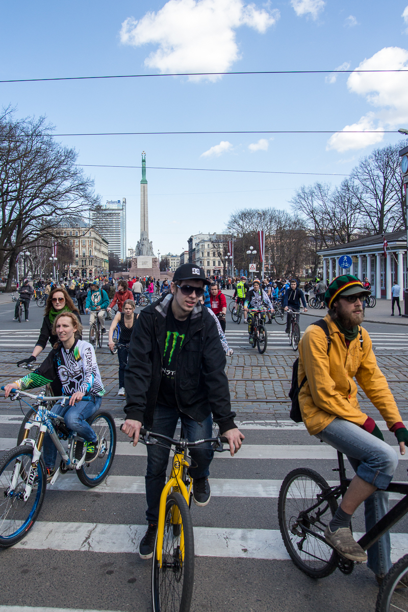 A large group of cyclists, including men and women of different ages, ride during the 2013 Kritiskā masa rally on a city street. In the background is a tall monument and buildings with modern and classic architecture. Trees without leaves line the street, and the sky is partly cloudy with blue patches.