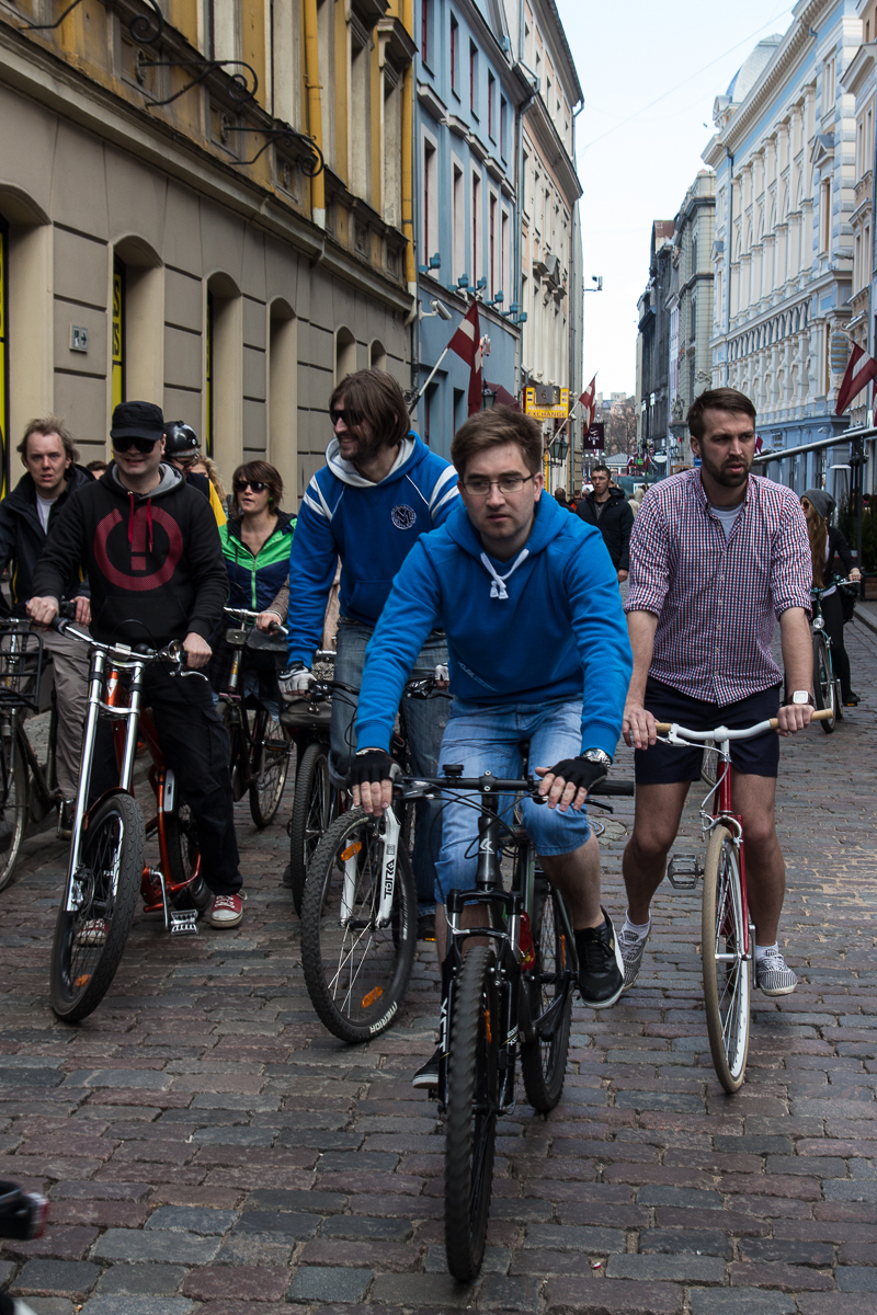 A group of people is riding bicycles on a cobblestone street in an urban area, reminiscent of the 2013 Kritiskā masa. The riders, wearing casual clothing including hoodies, jackets, and shorts, are focused ahead as they navigate the narrow, building-lined street. The background shows a mixture of colorful buildings and pedestrians.