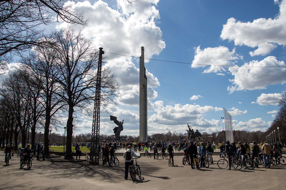 A large group of cyclists gather under a clear, sunny sky for the 2013 Kritiskā masa. Bare trees line the sides of the path, and statues flank a tall, narrow monument in the center distance. Puffy clouds float in the blue sky, creating a backdrop for the lively scene of people and bicycles.