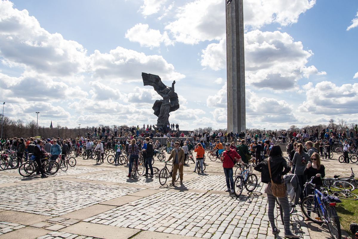 A large group of people, many with bicycles, gather at a public square on a partly cloudy day during the 2013 Kritiskā masa. Dominating the background is a tall, vertical monument and a dynamic sculpture of a figure holding a flag. The area is paved with light-colored stones and bricks.