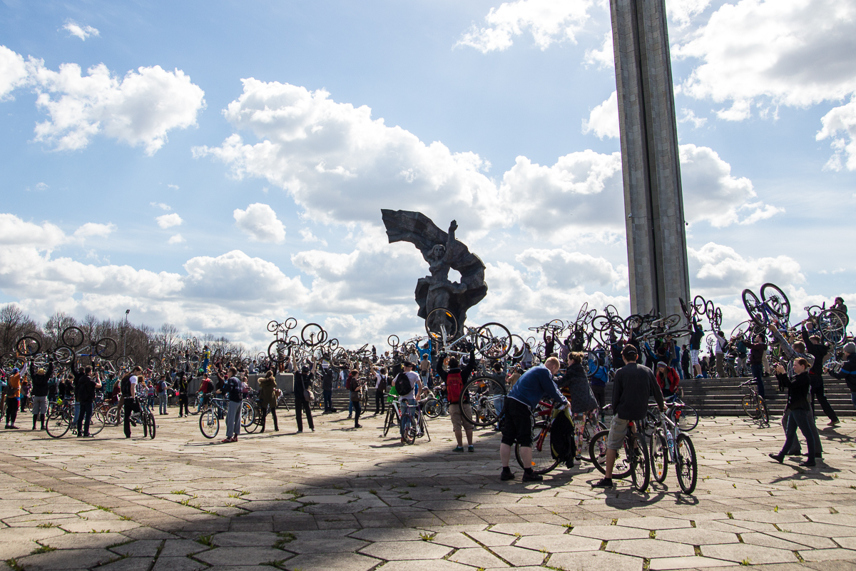 A large group of cyclists gather in a plaza on a sunny day, lifting their bicycles in the air in front of a towering sculpture. The sky is dotted with clouds, and many of the participants are dressed in casual clothing. This 2013 event marks Kritiskā masa, held on the stone-tiled plaza with a raised platform in the background.