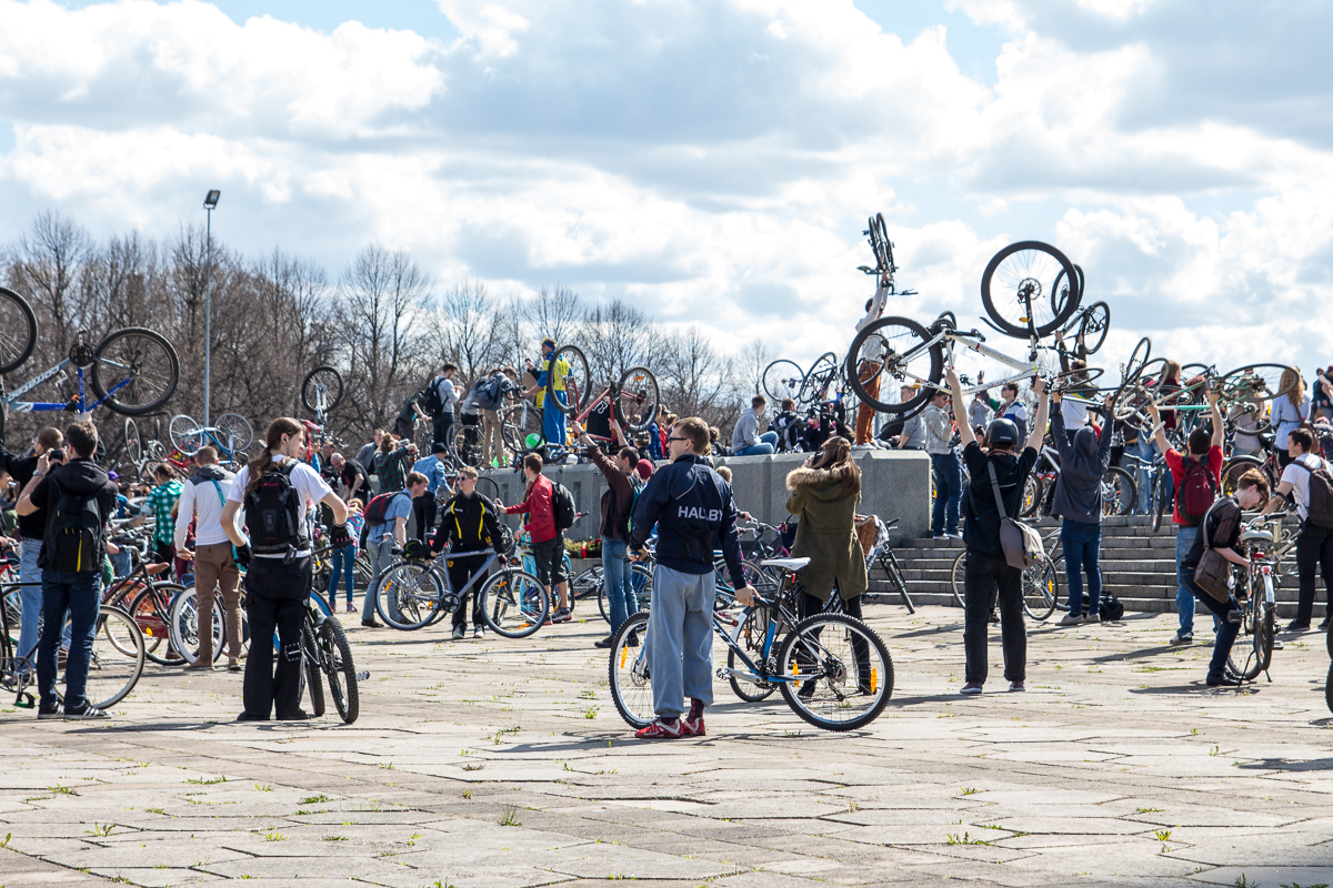 A large group of people gathered outdoors, each lifting their bicycles above their heads in celebration. The sky is partly cloudy, and leafless trees are visible in the background. The crowd spans a wide open area with paved ground, capturing the festive and communal spirit of Kritiskā masa 2013.