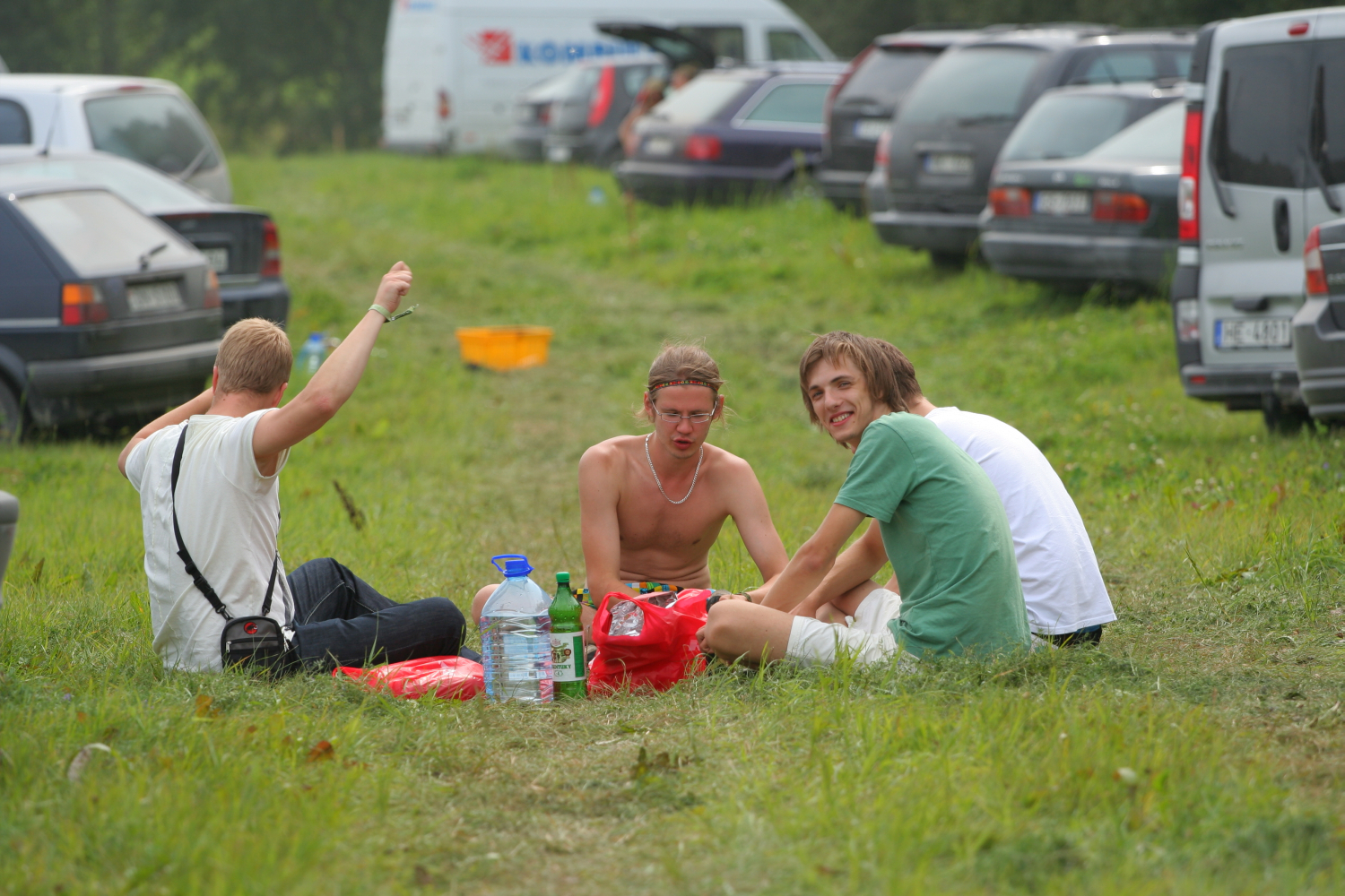 Four young men sit on the grass in a parking area between rows of parked cars, reminiscent of a 2011 summer gathering. They are gathered in a circle with some belongings, including plastic bottles and bags, in the middle. One of them, shirtless, smiles directly at the camera while another, wearing a green shirt, also smiles.