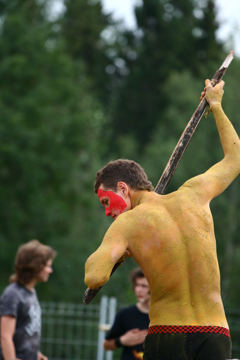 A person with their back to the camera is covered in yellow body paint with red paint on their face. They are holding a large stick above their head, poised to strike downward. The backdrop of blurred figures and green foliage hints at an outdoor setting, possibly from a 2011 Laba Daba festival.