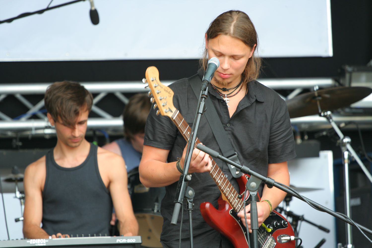 Two male musicians performing on stage. The foreground musician, with long brown hair, a black shirt, and playing a red electric guitar while singing into a microphone, exudes the rock vibes of 2011. The background musician has short dark hair, a black tank top, and is playing a Laba keyboard. Drums are partially visible.
