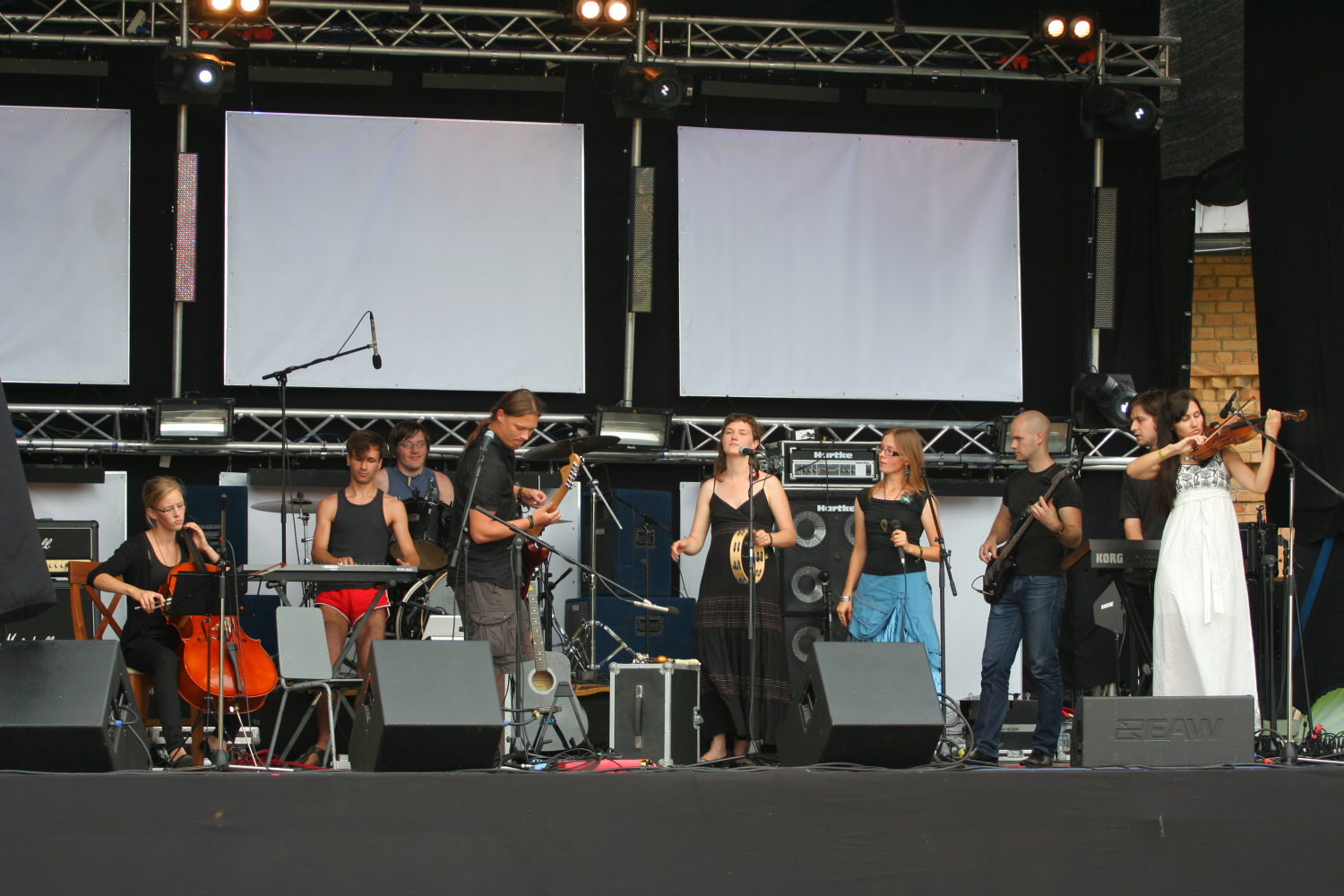 A band performing on an outdoor stage features nine musicians playing instruments including cello, violin, electric guitar, and bass. Some musicians are seated while others stand. They are surrounded by equipment such as microphones, stands, and speakers, with blank projection screens in the background. Laba daba 2011 vibes!