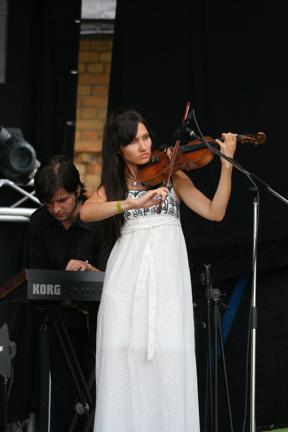A woman in a white dress plays a violin on stage, concentrating on her music. Behind her, a man dressed in black plays a Korg keyboard. The dark background reveals stage equipment and a brick wall. Both musicians appear focused on their performance at the 2011 Laba Daba festival.