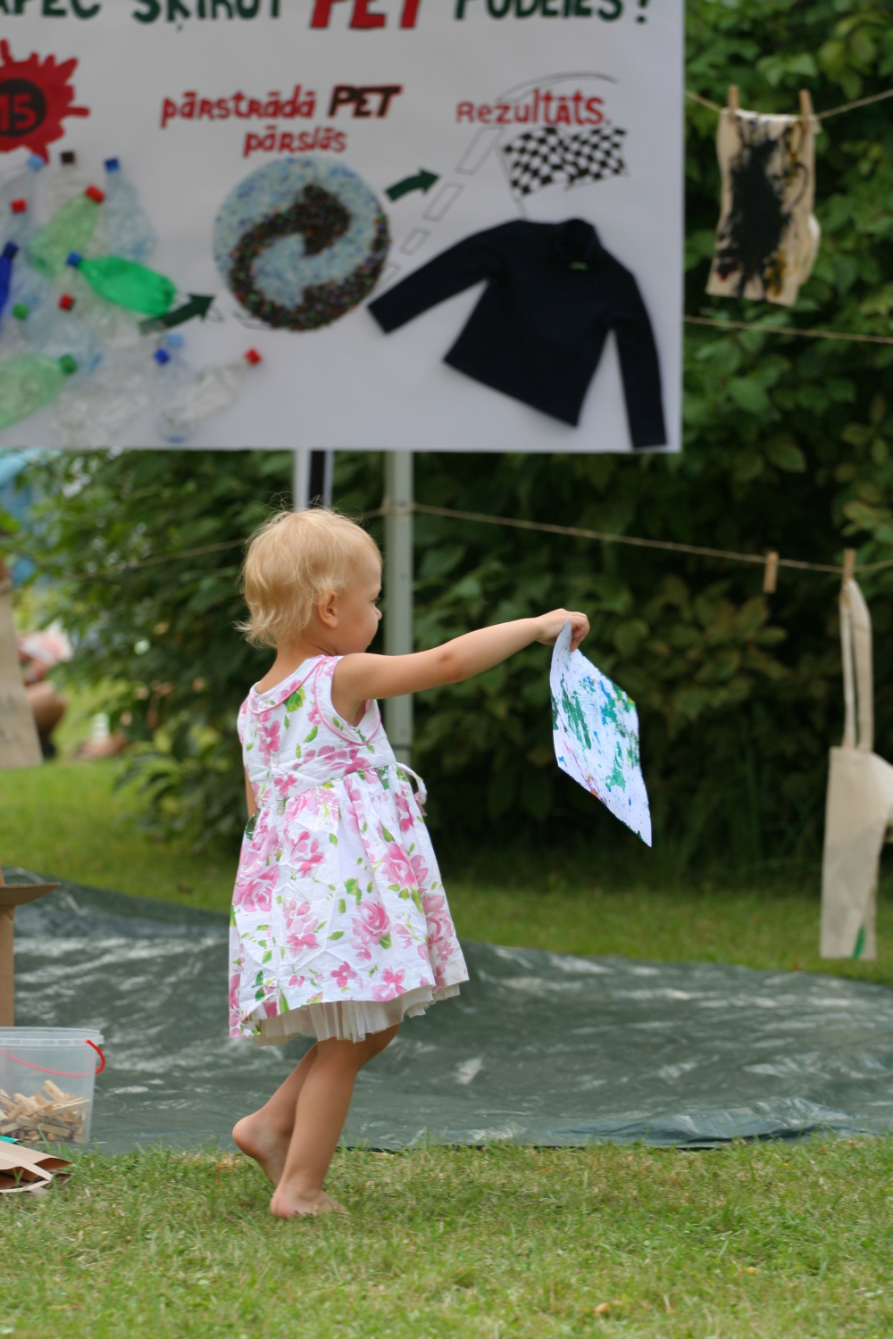 A young child with blonde hair, wearing a white dress with pink flowers, stands on grass and holds up a colorful drawing. Behind her, a sign with Latvian text and a collage are visible. The scene appears to be an outdoor daba event or activity in a garden or park with trees and greenery in the background.