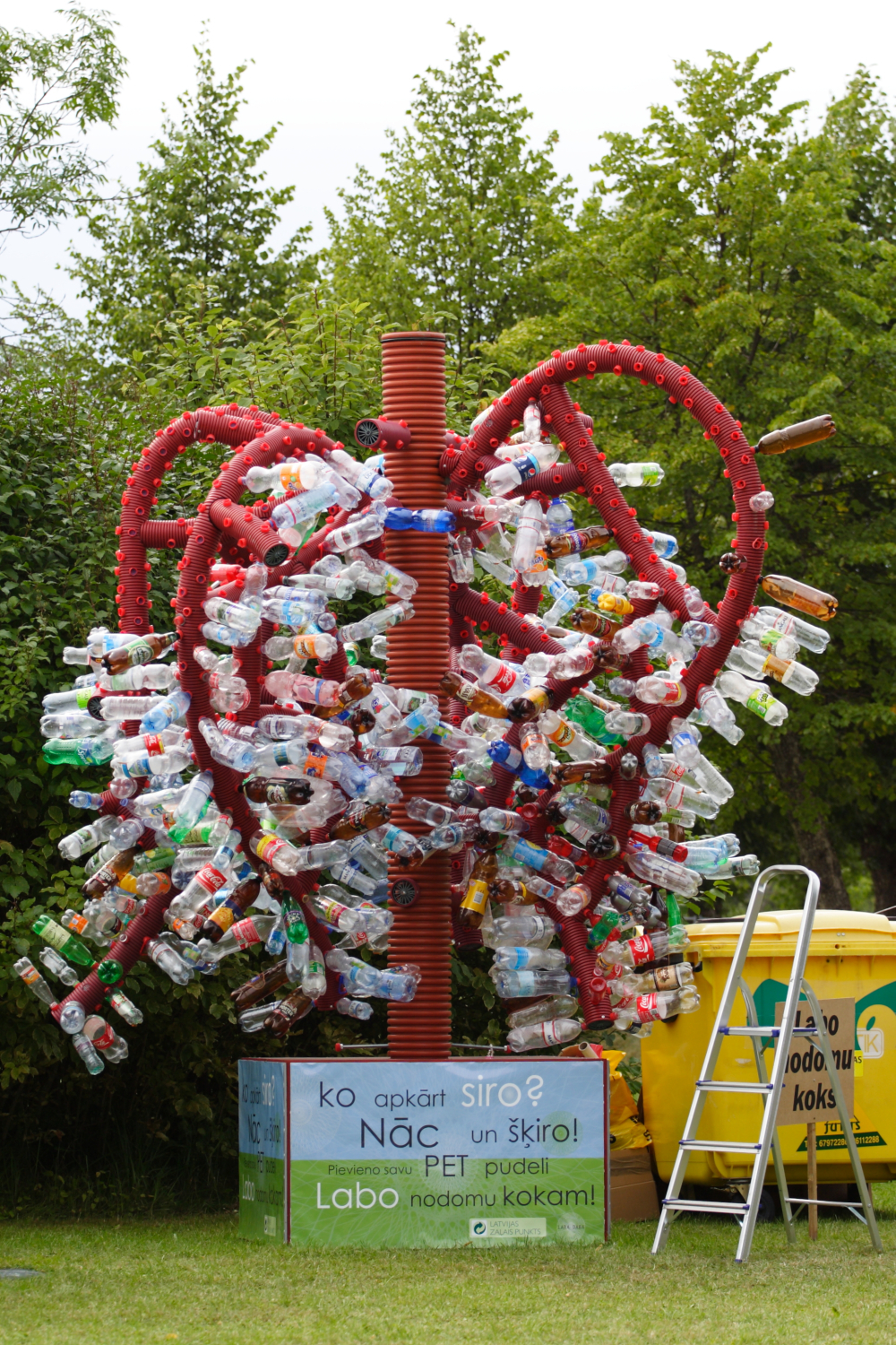 A red metal sculpture in the shape of a tree, created for the 2011 Laba Daba festival, holds numerous attached plastic bottles. Positioned outdoors with lush green trees in the background, it stands next to a yellow recycling bin and a silver ladder, with a sign at its base encouraging plastic bottle recycling in Latvian.
