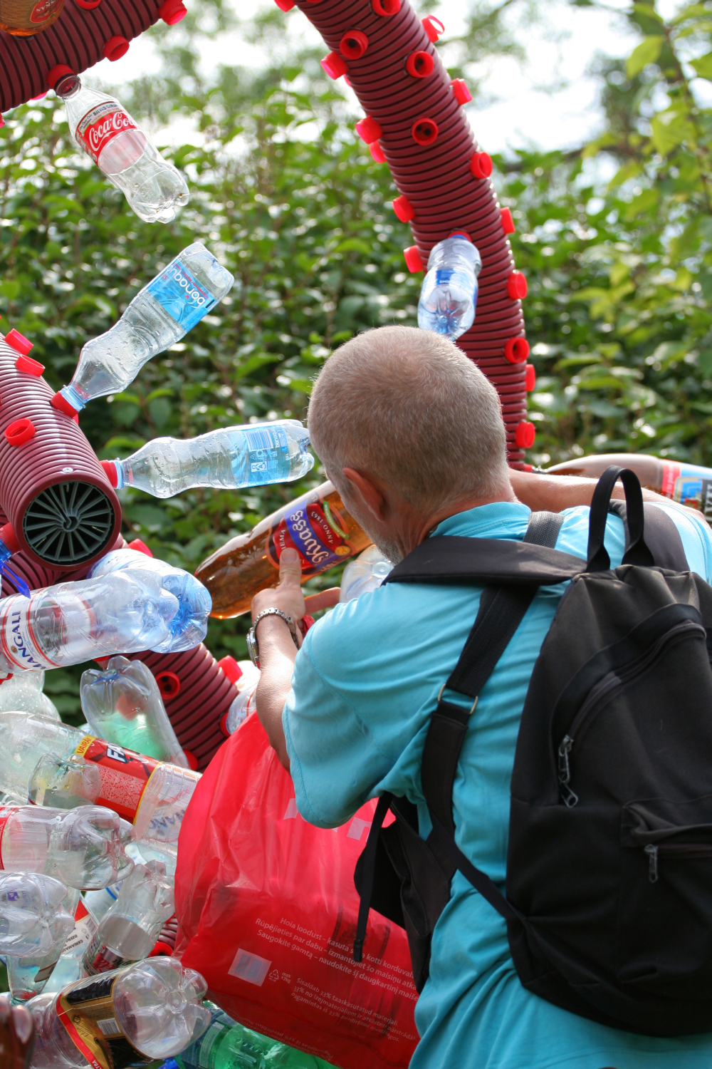 A person with cropped hair, wearing a turquoise shirt and carrying a black backpack, places a plastic bottle into a recycling sculpture made of numerous plastic bottles and other items at the Laba Daba 2011 festival. The background features green foliage and blurred outdoor scenery.