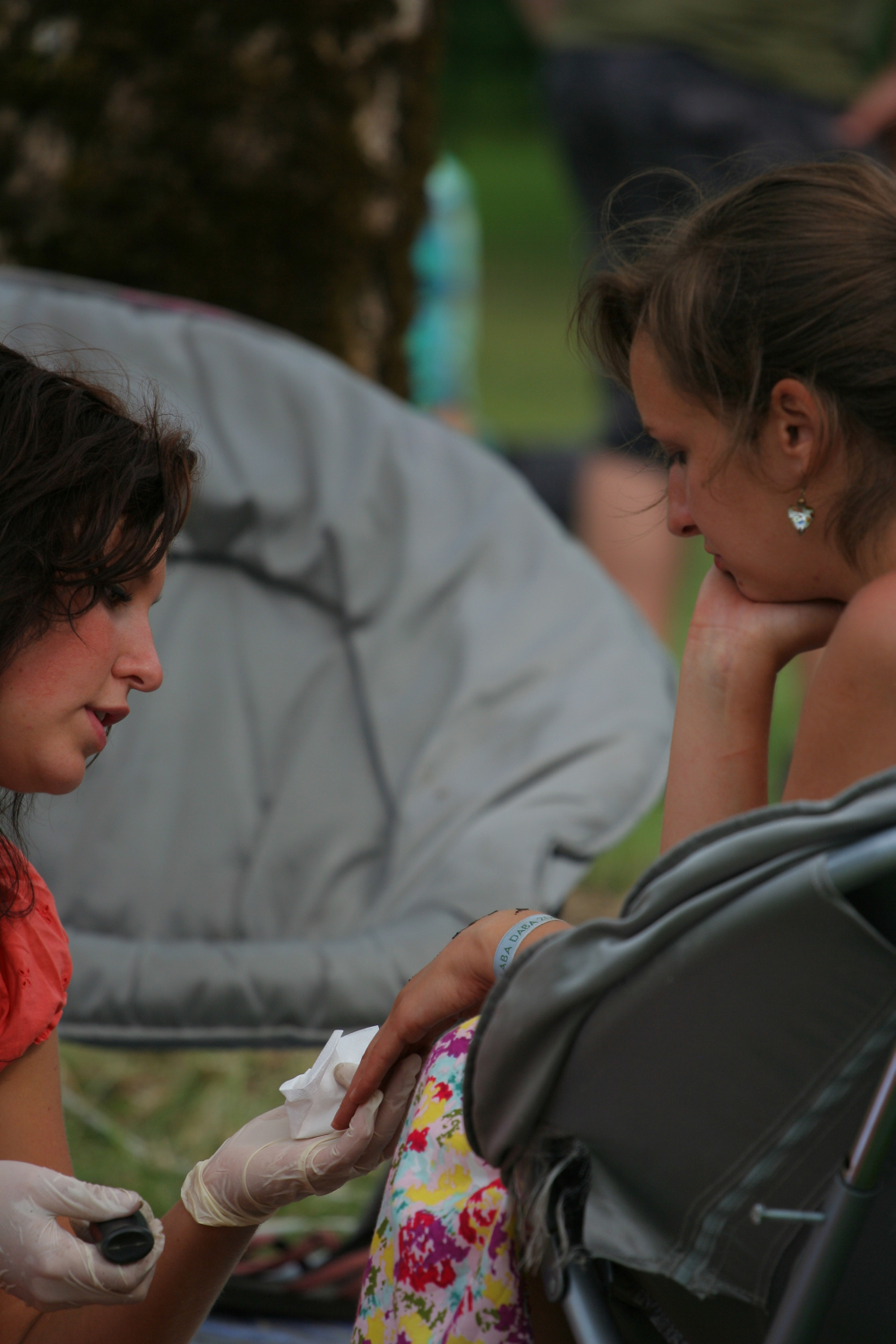Two women are seated outdoors on foldable chairs. One woman, wearing an orange shirt and white gloves, is carefully applying a bandage to the other woman's hand, who is wearing a colorful floral dress from 2011. Both are deeply focused on the task. The background shows a grassy area and blurred people.