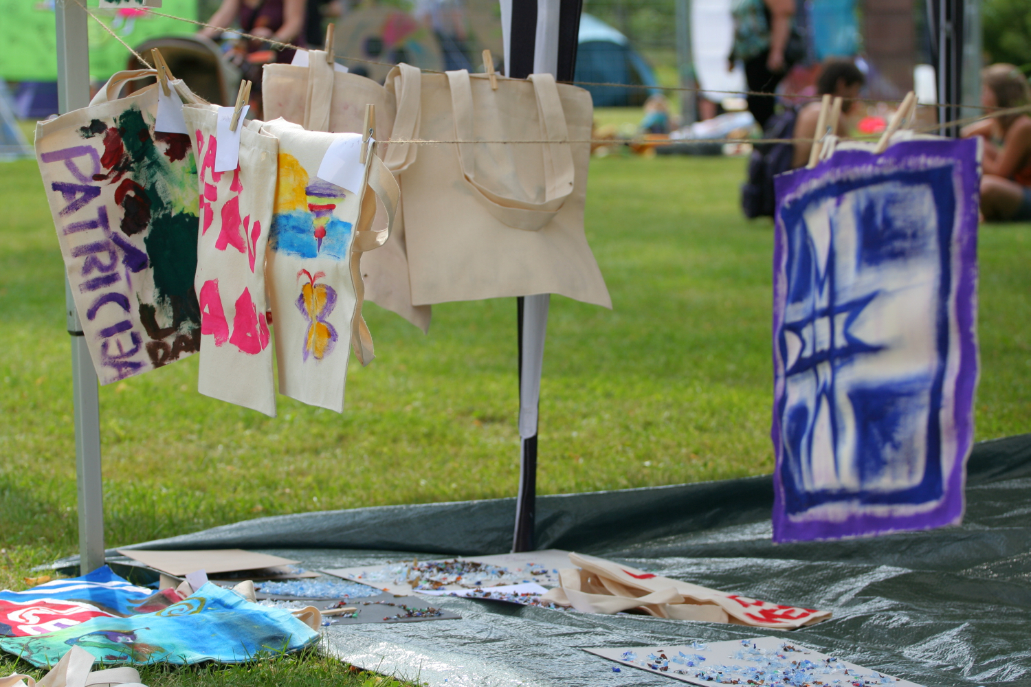Fabric tote bags with colorful hand-painted designs, including abstract patterns and a butterfly, hang to dry on a clothesline at the 2011 Laba Daba outdoor arts and crafts fair. A green grassy field and people seated in the background are slightly blurred. Some art supplies are scattered on the ground below.