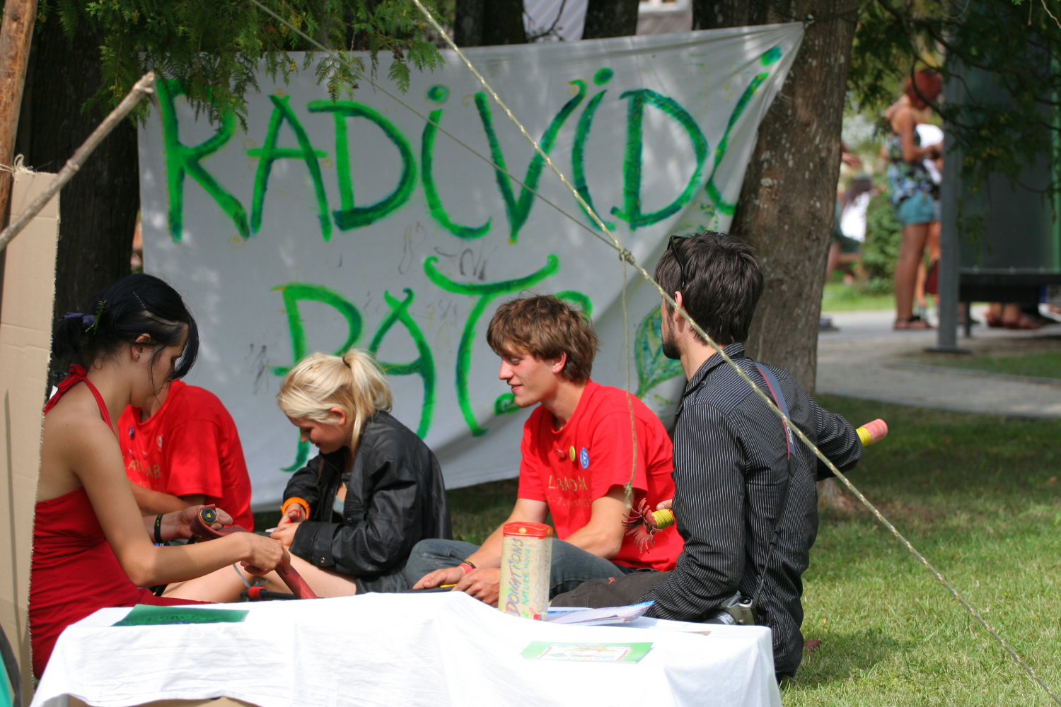 A group of four young individuals sits on the grass in a park, creating art. Three wear red shirts, and one is in black. Behind them, a banner reads "RAD I VIDI PATOM." They use materials like paper and paints. Trees and other people are visible in the background, evoking a nostalgic Laba daba vibe from 2011.