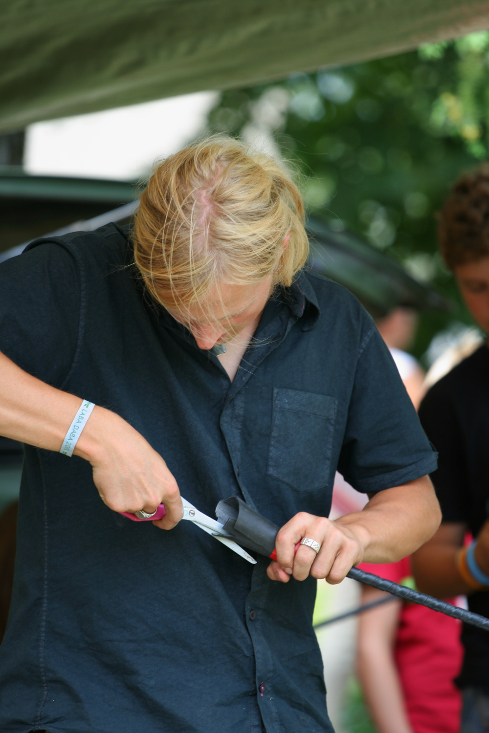 A person with long blond hair wearing a black shirt is focused on cutting black material with a pair of scissors. They are outdoors, under a canopy in what appears to be a 2011 Laba Daba festival, and other people are in the background. The person is wearing a light blue wristband and a ring on their right hand.
