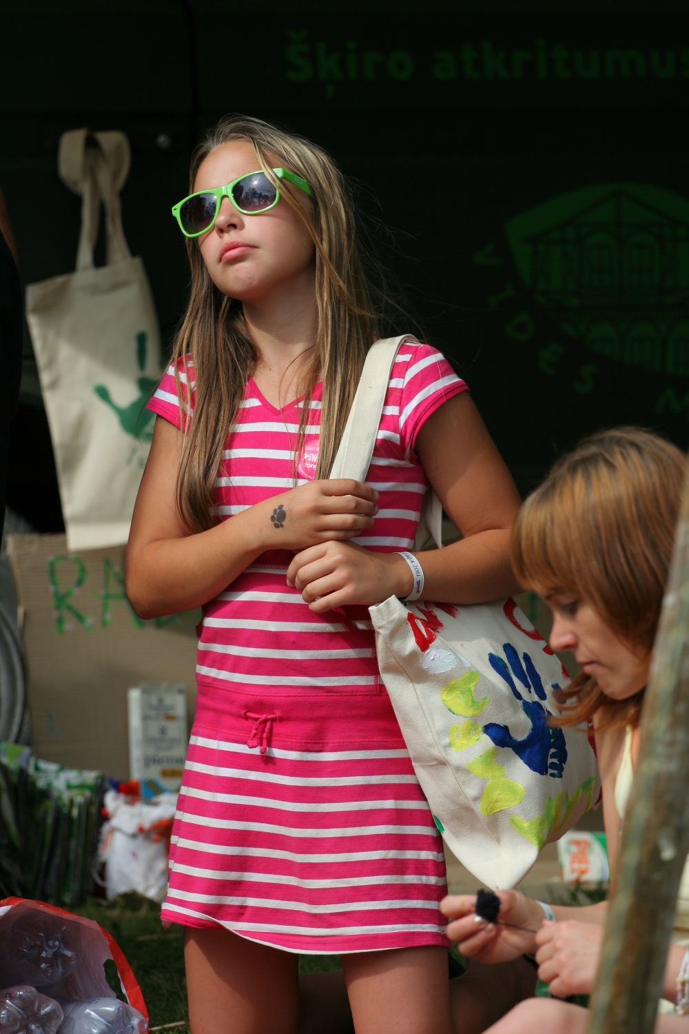 A young girl wearing a pink and white striped dress and green sunglasses is holding a tote bag decorated with colorful handprints. She stands at an outdoor Laba daba event with a focused expression. Another person, partially visible, is seated nearby, engaged in an activity.