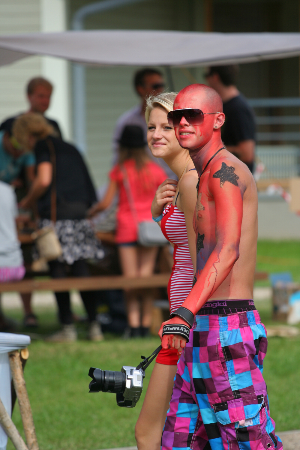 A man and woman walk outdoors, smiling. The man, shirtless and painted red with a black star on his chest, wears sunglasses and colorful checkered shorts from Laba daba 2011. He holds a camera. The woman wears a red-striped sleeveless shirt. Other people and picnic tables are blurred in the background.
