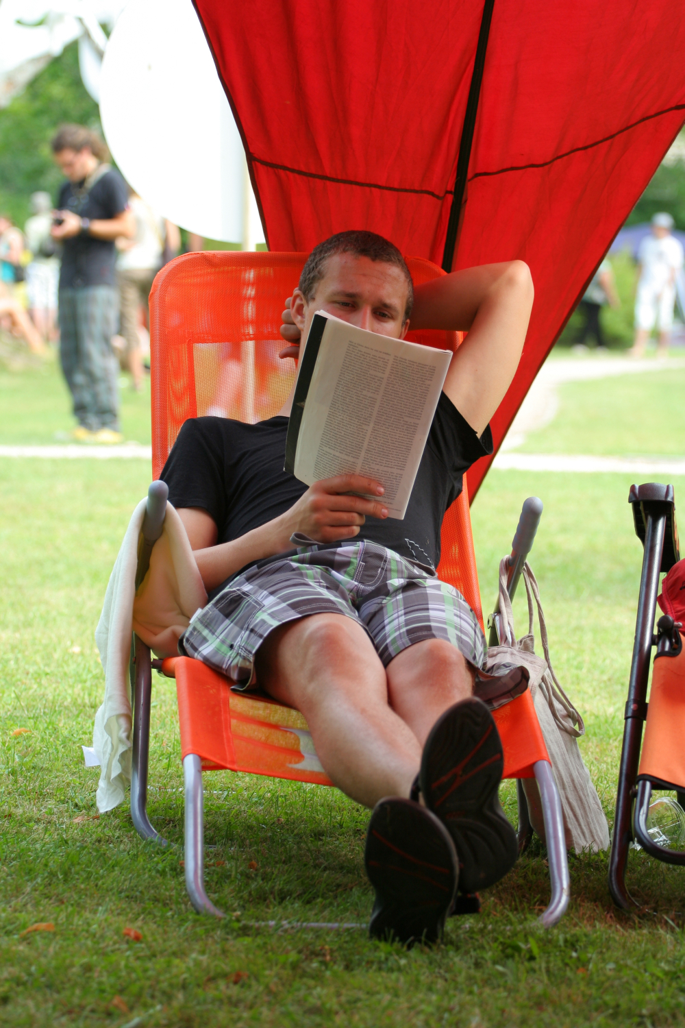 A man relaxes on an orange lounge chair with a red canopy, reading a book from 2011. He wears a black t-shirt and plaid shorts, with one arm resting behind his head. His legs are crossed, and his sandals are touching the grass. The scene is outdoors, with blurred people and greenery in the background—laba daba.