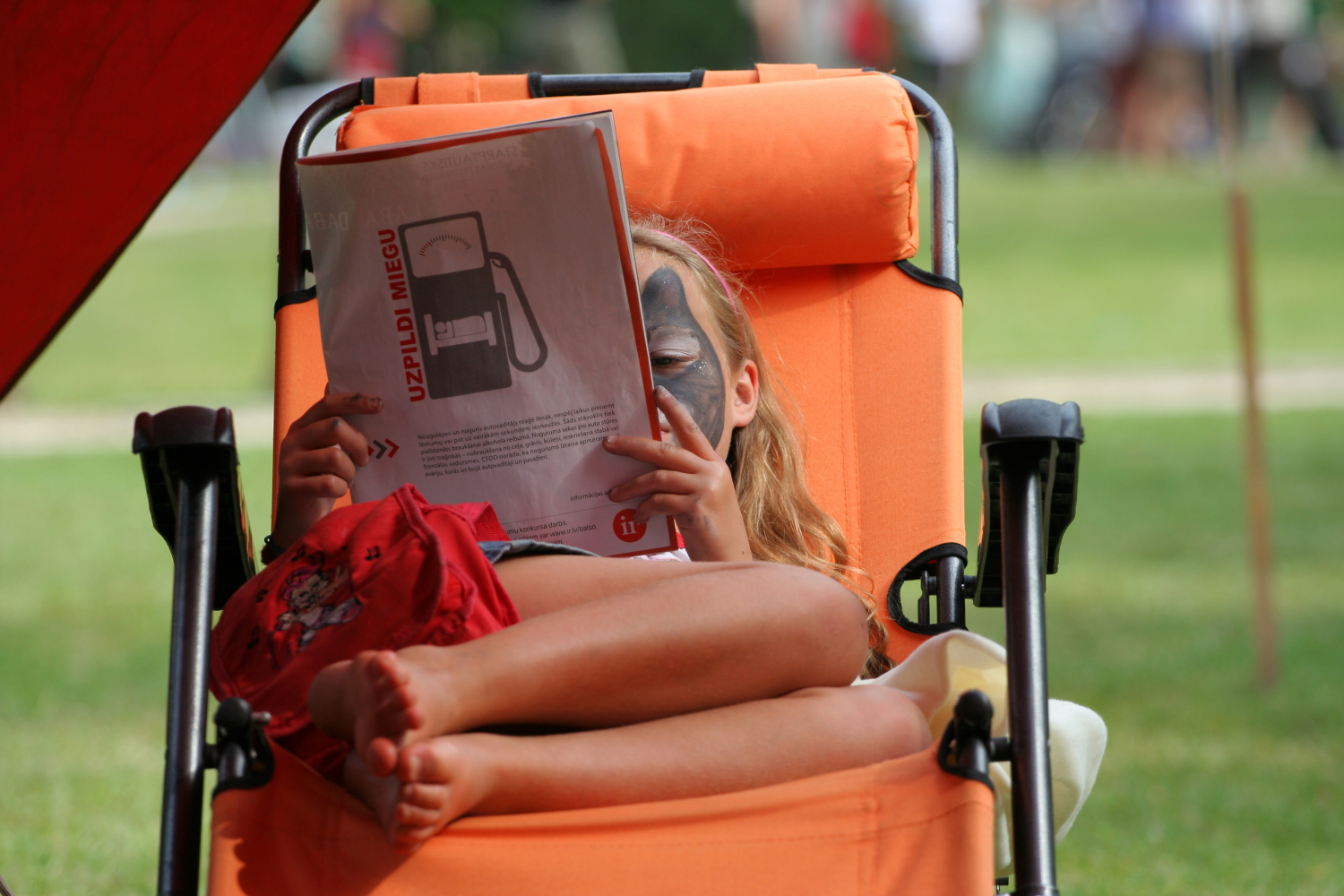 A child with face paint lies on an orange chair outdoors, reading a 2011 magazine. The cover features a gasoline pump and text in a non-English language. The child's legs are crossed and relaxed, while the background shows grassy area and blurred figures, creating a tranquil Laba daba scene.