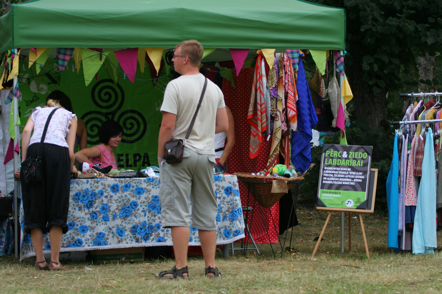 A man in a white shirt and beige shorts stands in front of a green tent with colorful triangular flags, reminiscent of Laba Daba 2011. Two women behind a table with a blue floral cloth are selling goods. To the right, a rack of clothes is displayed. A nearby sign contains text, and a leafy tree is in the background.