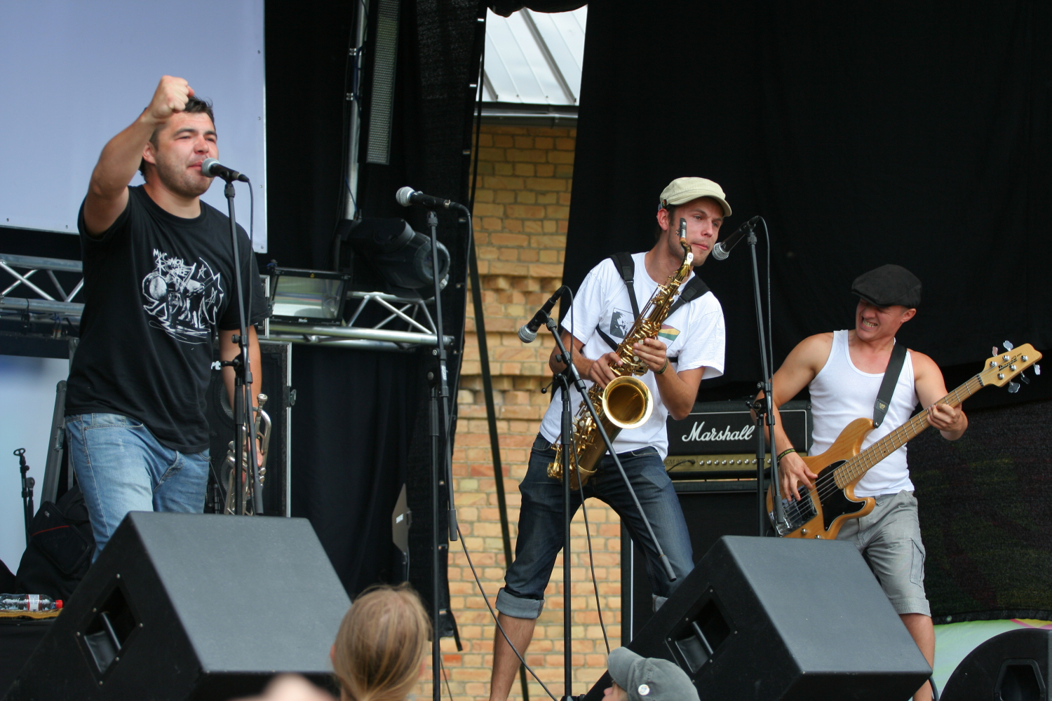 Three male musicians perform on an outdoor stage. The first sings into a microphone, the second plays a saxophone, and the third plays a bass guitar. A crowd watches them near two speakers at the front of the stage. The backdrop includes a brick wall and various sound equipment from 2011, adding to the Laba-daba vibe.