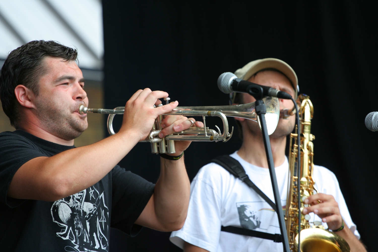 Two musicians perform on stage. The musician on the left plays a trumpet, wearing a black t-shirt with white graphics, reminiscent of 2011 styles. The musician on the right, in a white t-shirt, sunglasses, beige cap, and microphone headset, skillfully plays a saxophone. Both are set against a dark background.