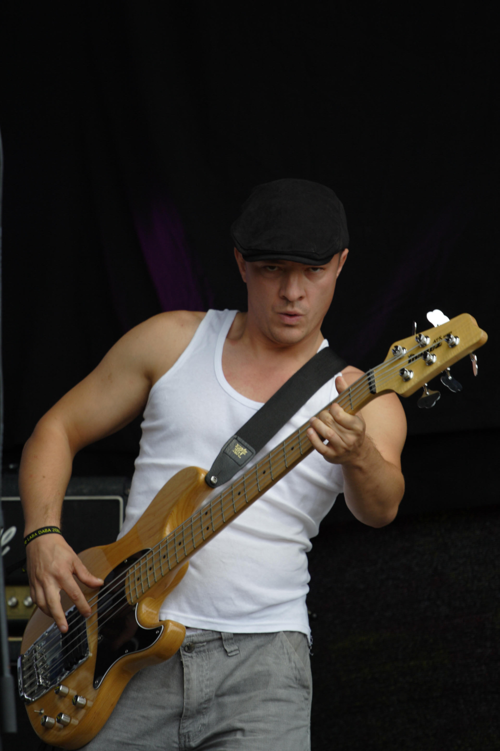 A musician in a white tank top and gray pants plays an electric bass guitar, reminiscent of the 2011 Laba style. He wears a black flat cap and a serious expression. His left hand holds the guitar's neck, while his right hand plucks the strings. A dark background and an amplifier are visible behind him.