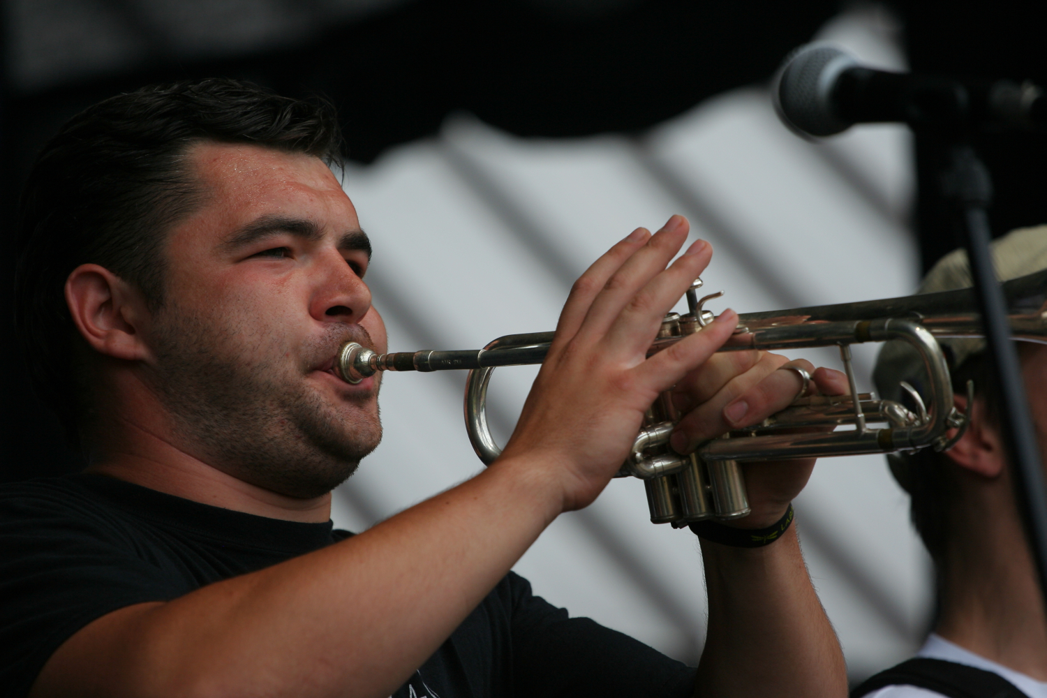 A man wearing a black shirt plays a silver Laba trumpet with focus and intensity. He stands in front of a microphone, captured mid-performance. The background is blurred, emphasizing the foreground subject. The lighting highlights his concentrated expression and the shiny surface of the trumpet.
