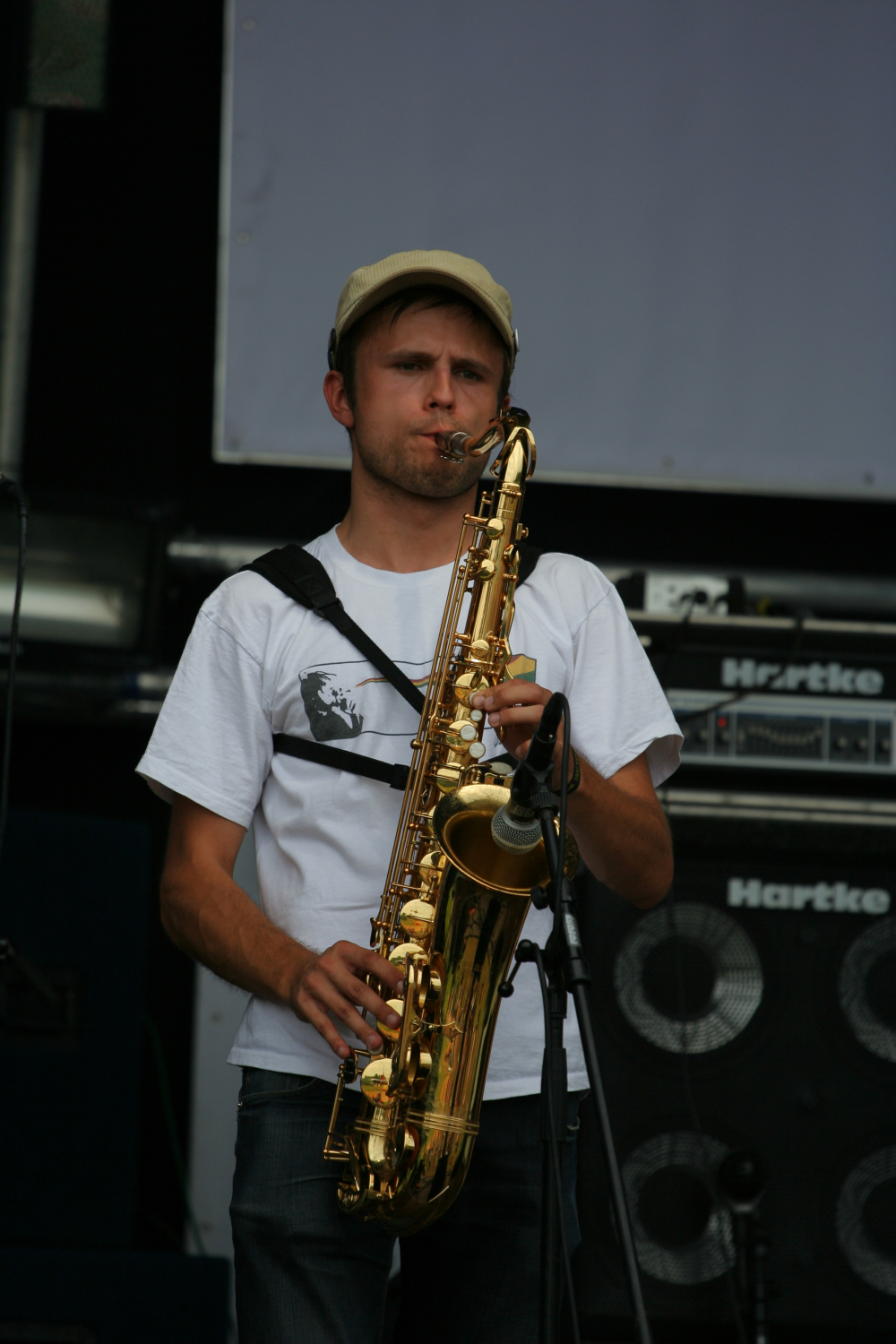 A man wearing a beige cap and a white t-shirt plays a saxophone on stage. He stands in front of a Hartke amplifier stack, focusing intently on his performance. A microphone captures the sound near the saxophone, with additional stage equipment visible in the background. The scene evokes memories of 2011 Laba daba performances.