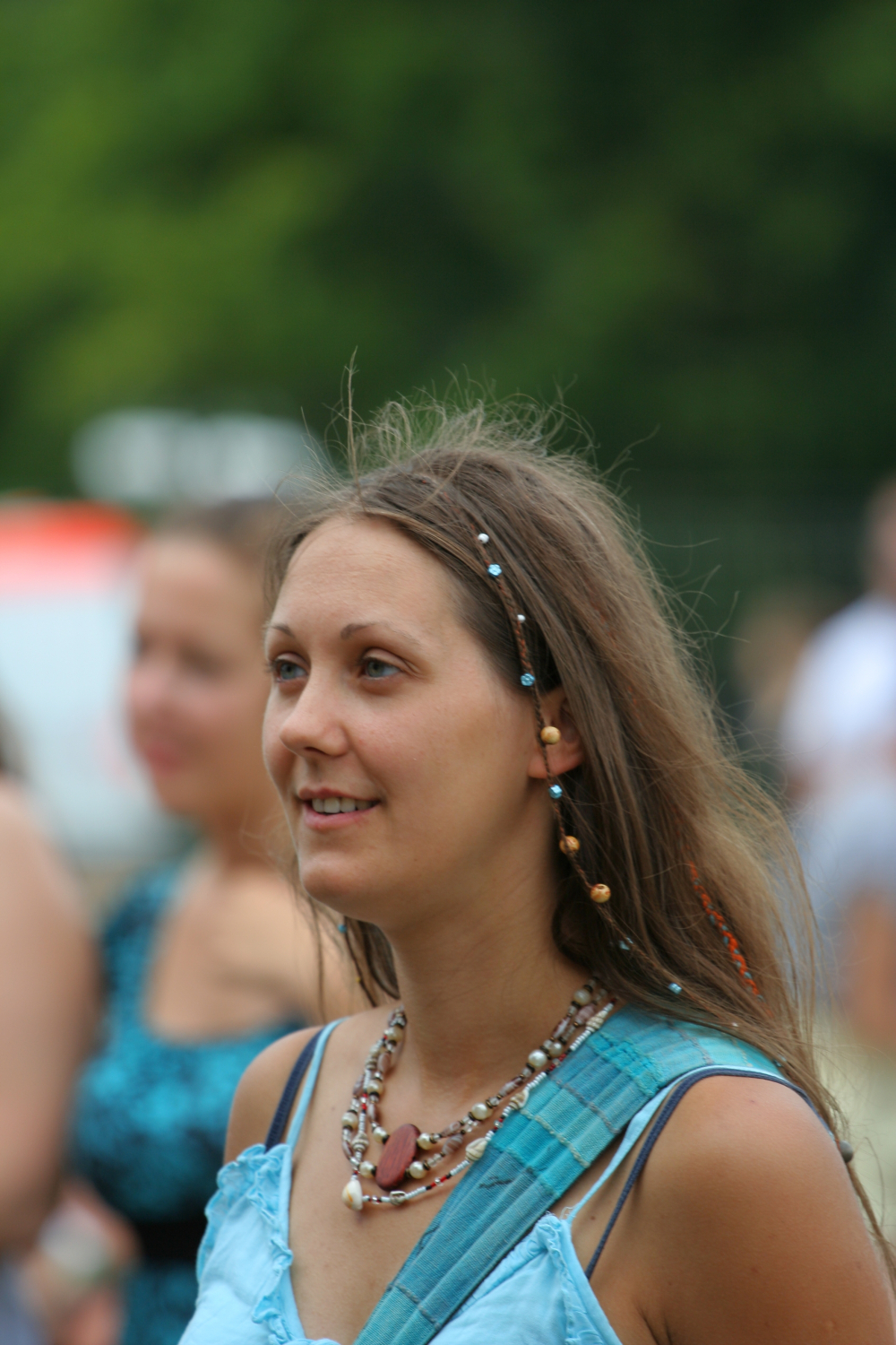 A woman with long brown hair adorned with beads smiles while looking to the side. She's wearing a light blue sleeveless top and layered necklaces with various beads and a large red pendant, reminiscent of laid-back 2011 Laba daba vibes. The background shows slightly blurred people and greenery, indicating an outdoor setting.