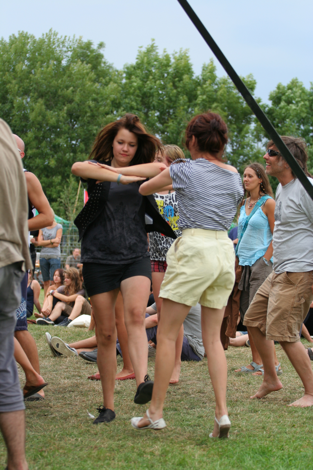 Two women dance energetically in a grassy outdoor festival at Laba Daba 2011. One wears a black top and skirt, and the other a striped shirt with white shorts. People sit and stand in the background, enjoying the event. Trees and a cloudy sky are visible, creating a lively summer atmosphere.