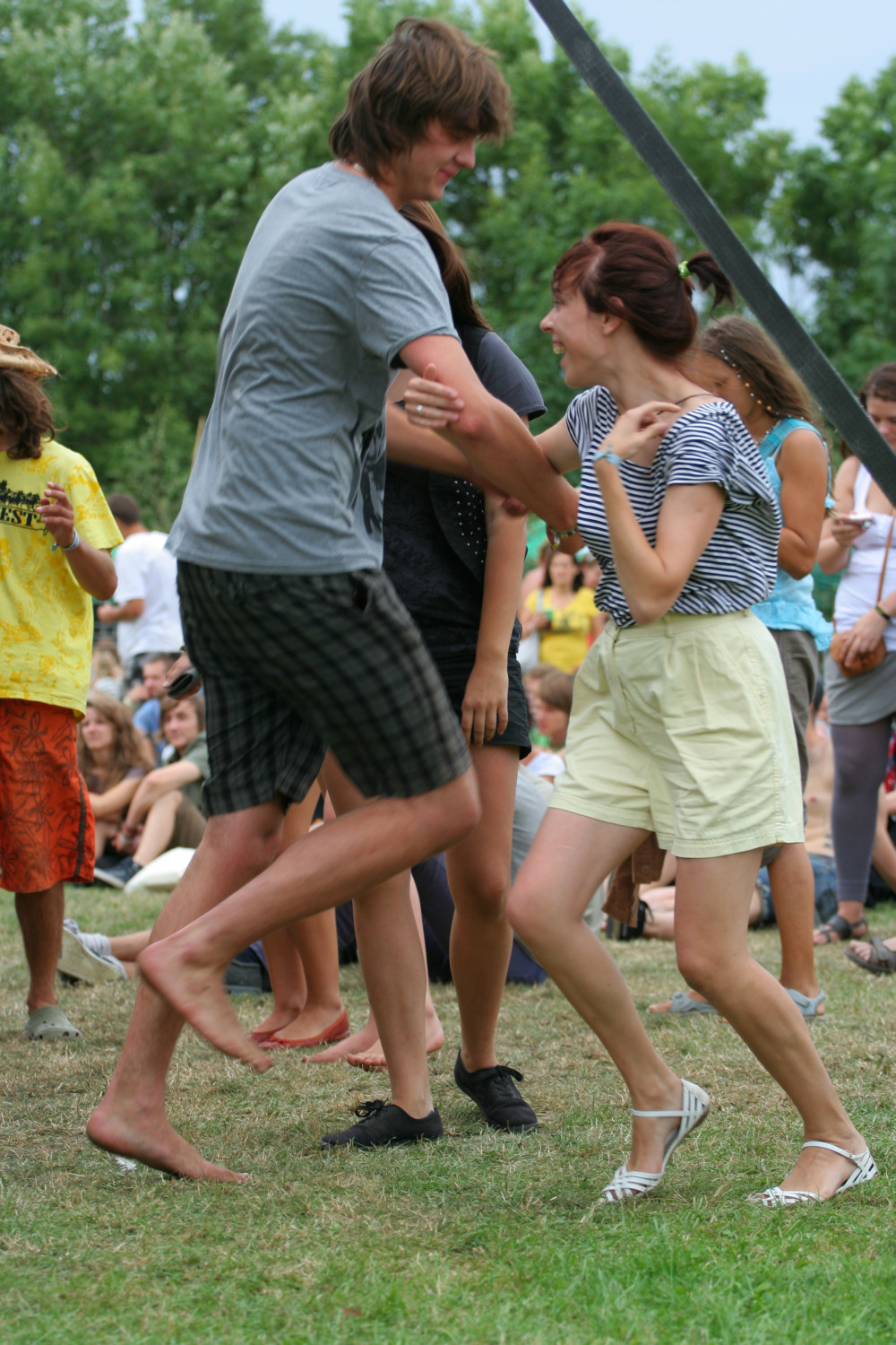 A group of young people are dancing and having fun outdoors on a grassy area. A young man in a gray T-shirt and plaid shorts is enthusiastically dancing with a young woman in a striped shirt and light-colored shorts. It's 2011, and other people in casual summer clothes are watching and enjoying the scene under the trees.