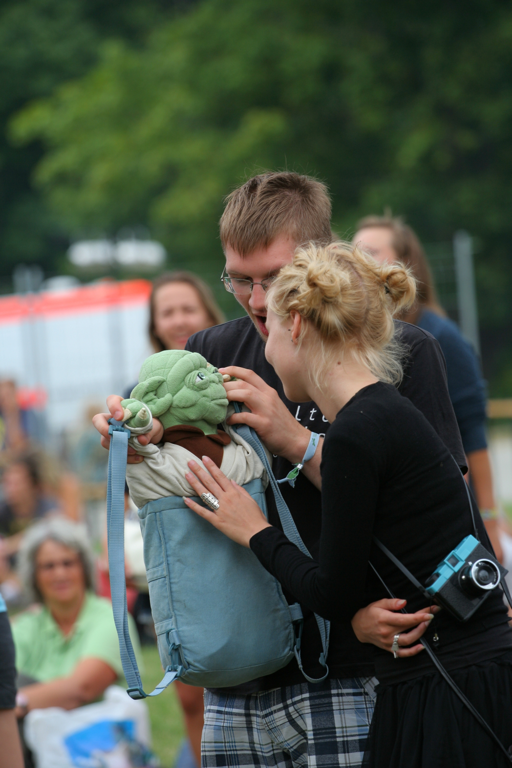A man and woman are outdoors holding a plush toy inside a blue backpack. The toy resembles Yoda from Star Wars. The woman, wearing a black outfit with a camera around her neck, is smiling while adjusting the toy. The man, in glasses and a black shirt from Laba's 2011 collection, is also focused on the toy. People and trees are blurred in the background.