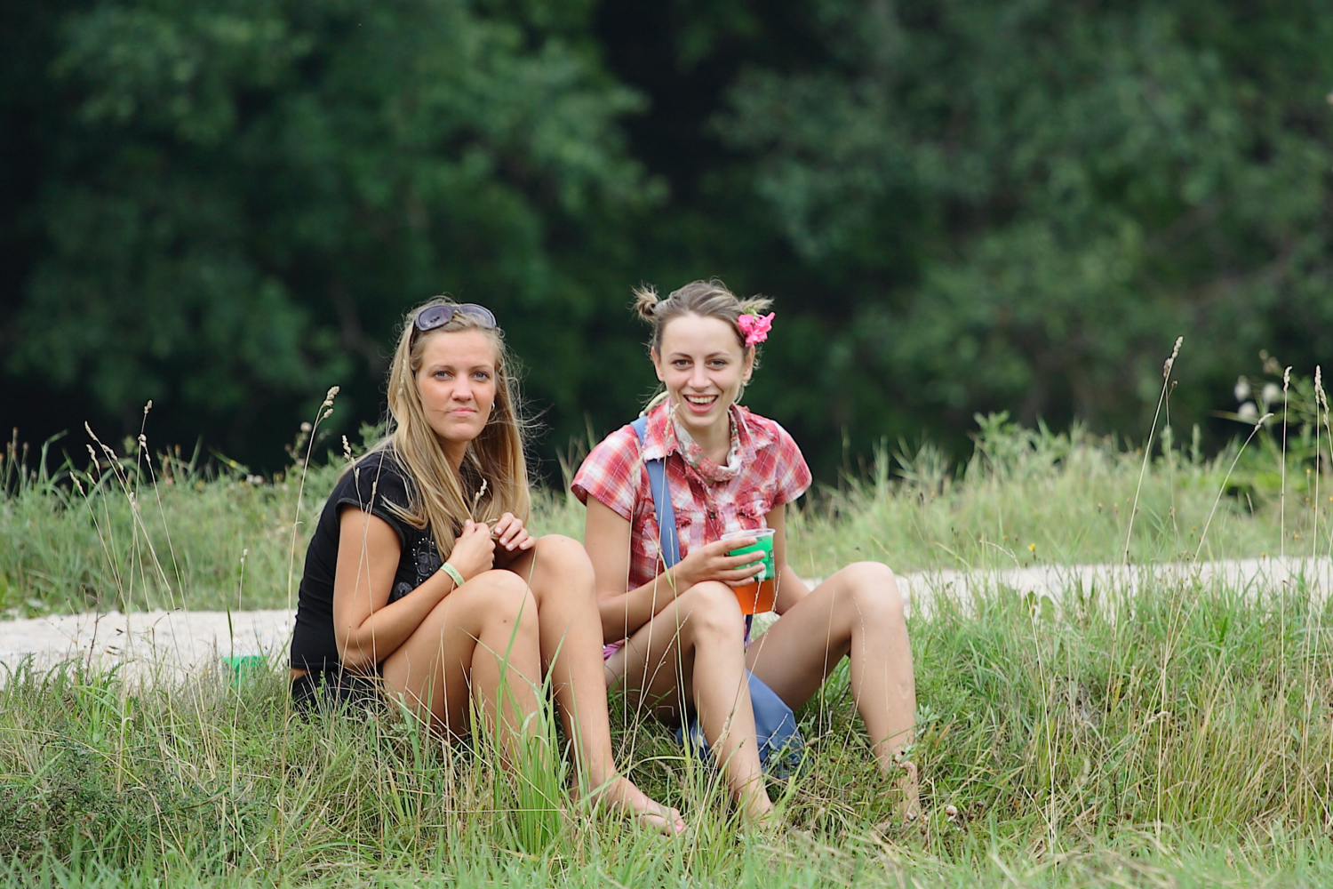 Two women sit on grass in casual summer attire at Laba Daba 2011. The woman on the left wears a black shirt and sunglasses atop her head, while the woman on the right, smiling, dons a red plaid shirt and has flowers in her hair. They are surrounded by lush greenery, with trees in the background and long grass in the foreground.