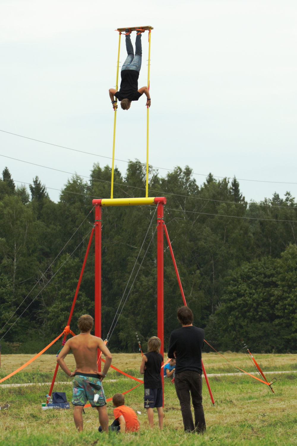 An individual is balancing upside down on a horizontal bar, suspended high above the ground on a tall structure with yellow and red components, while four onlookers watch from below in an open field with trees in the background. The scene captures the daring spirit of Laba Daba 2011, an outdoor event filled with excitement.