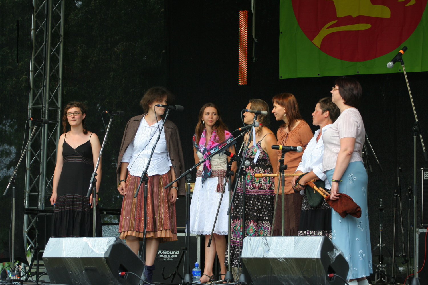 A group of six women stand on a stage in front of microphones, preparing to sing. They are dressed in a mix of casual and traditional clothing, reminiscent of 2011 fashion. There is a green banner with a red and yellow design hanging behind them. Musical equipment, such as speakers and a drum set, is visible on stage.
