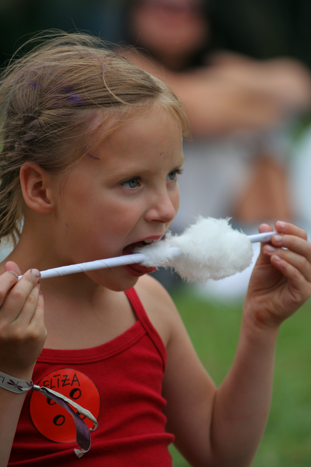 A young girl wearing a red tank top is enjoying cotton candy at Laba Daba 2011. She has light brown hair with a small purple streak and holds the cotton candy on a white stick. The girl sports a red sticker on her top and a gray wristband, while the blurred background shows other festival-goers.