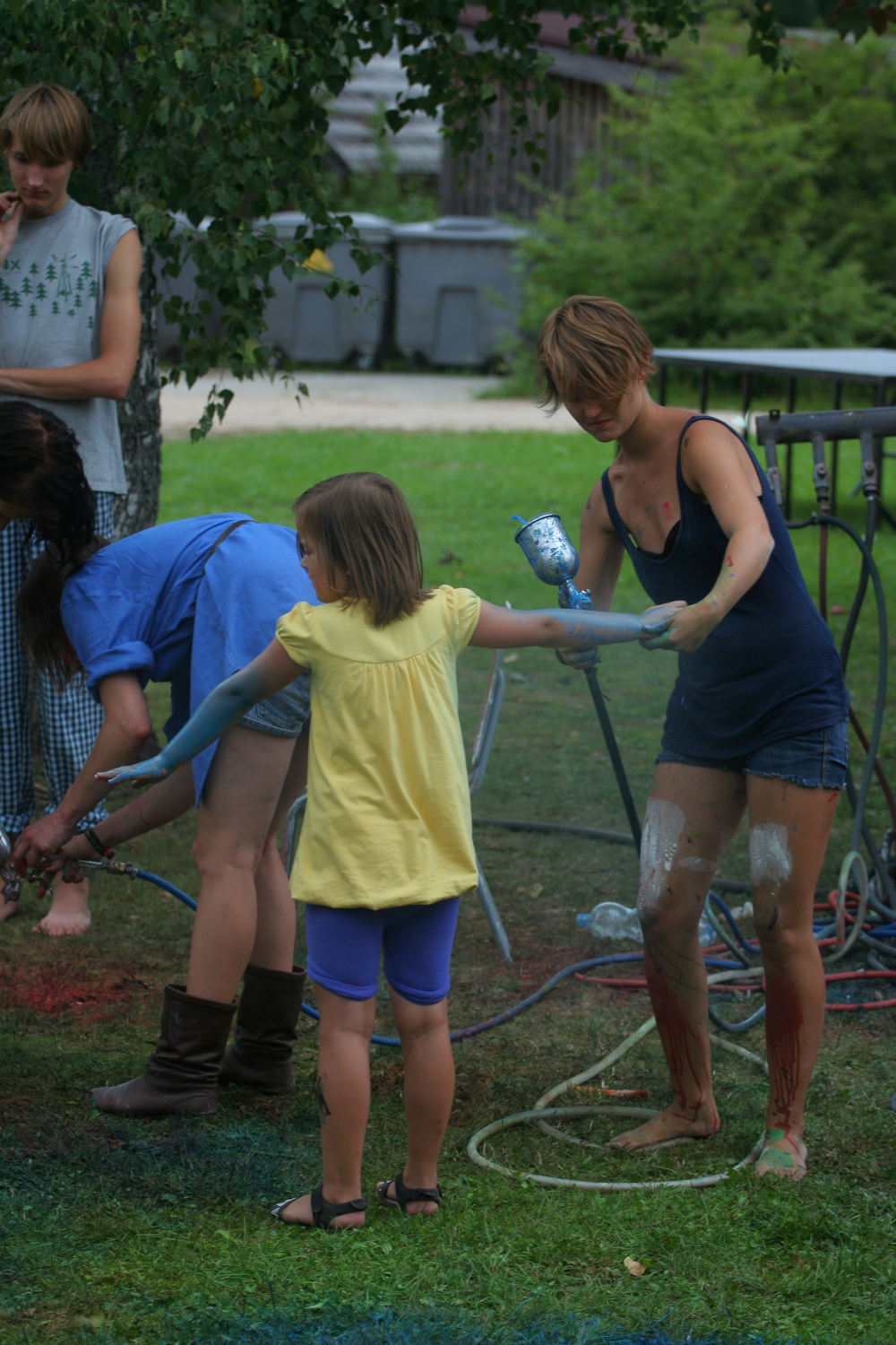 A young girl in a yellow shirt and blue shorts holds hands with a woman in a black tank top and blue shorts. Both are covered in paint. Another woman in a blue dress and boots is bent over nearby. A man in a gray tank top stands in the background, enjoying the vibrant Laba daba festival of 2011 on the grassy area.