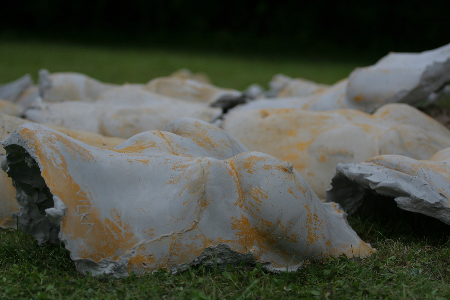 Broken stone or plaster sculptures of human torsos, partially covered in yellow and white paint, lie on green grass. The fragments are scattered, evoking a Laba daba vibe set against a dark green blurred background, suggesting an outdoor setting like a park or garden in 2011.